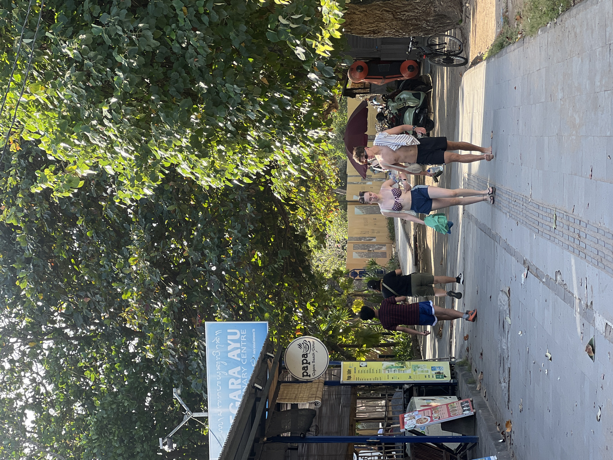 Two young adults, a male and a female, are walking down a paved sidewalk next to a line of parked motorbikes and scooters. The male is shirtless and wearing black shorts, while the female is wearing a polka dot bikini top and blue shorts. They are walking away from the camera, towards the right side of the image. In the background, there are trees and foliage, and the sun appears to be shining, casting shadows on the ground.
To the left of the sidewalk, there is a building with a sign that reads "GARA AYU PRIMARY CENTRE" in English, with some Balinese script above it. There is also a circular sign with "papa" and "Segara Beach Sanur" visible. A few other people are walking further down the sidewalk in the distance. The scene appears to be a street in a tropical location, likely during the daytime, with a relaxed atmosphere. The overall impression is one of a warm, sunny day in a tourist area.