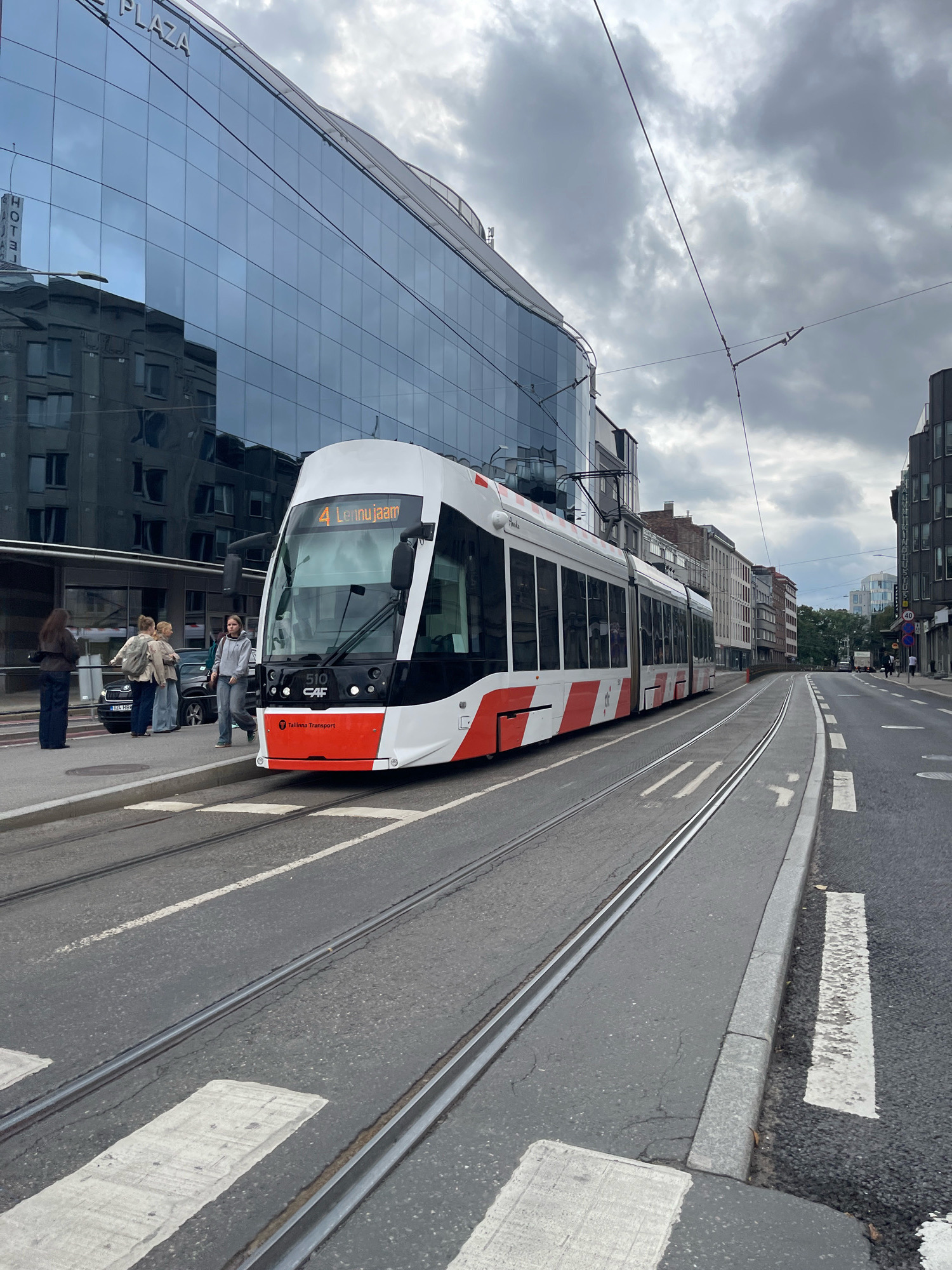 The image depicts a modern white and red tram with the number 4 and "Lennujaam" displayed on its front, indicating its route to the airport. The tram is stopped at a tram stop on a paved street in Tallinn, Estonia. A group of four individuals are standing at the tram stop, appearing to be waiting for the tram. One woman, wearing a gray hoodie and jeans, is in the foreground, looking towards the camera. Another woman, wearing a brown jacket and dark pants, is standing further back, facing away from the camera. Two other individuals, one with a backpack, are also at the stop.
The background features a large, modern building with a glass facade that reflects the sky and surrounding buildings. The sky is overcast with dramatic clouds. The street has tram tracks running through it, with a crosswalk visible in the foreground. The scene suggests an urban environment, likely a busy city street during daylight hours. The tram appears to be a recent model, and the building's architecture suggests contemporary urban development. The overall impression is that of a typical scene in a European city, with public transportation and modern architecture.