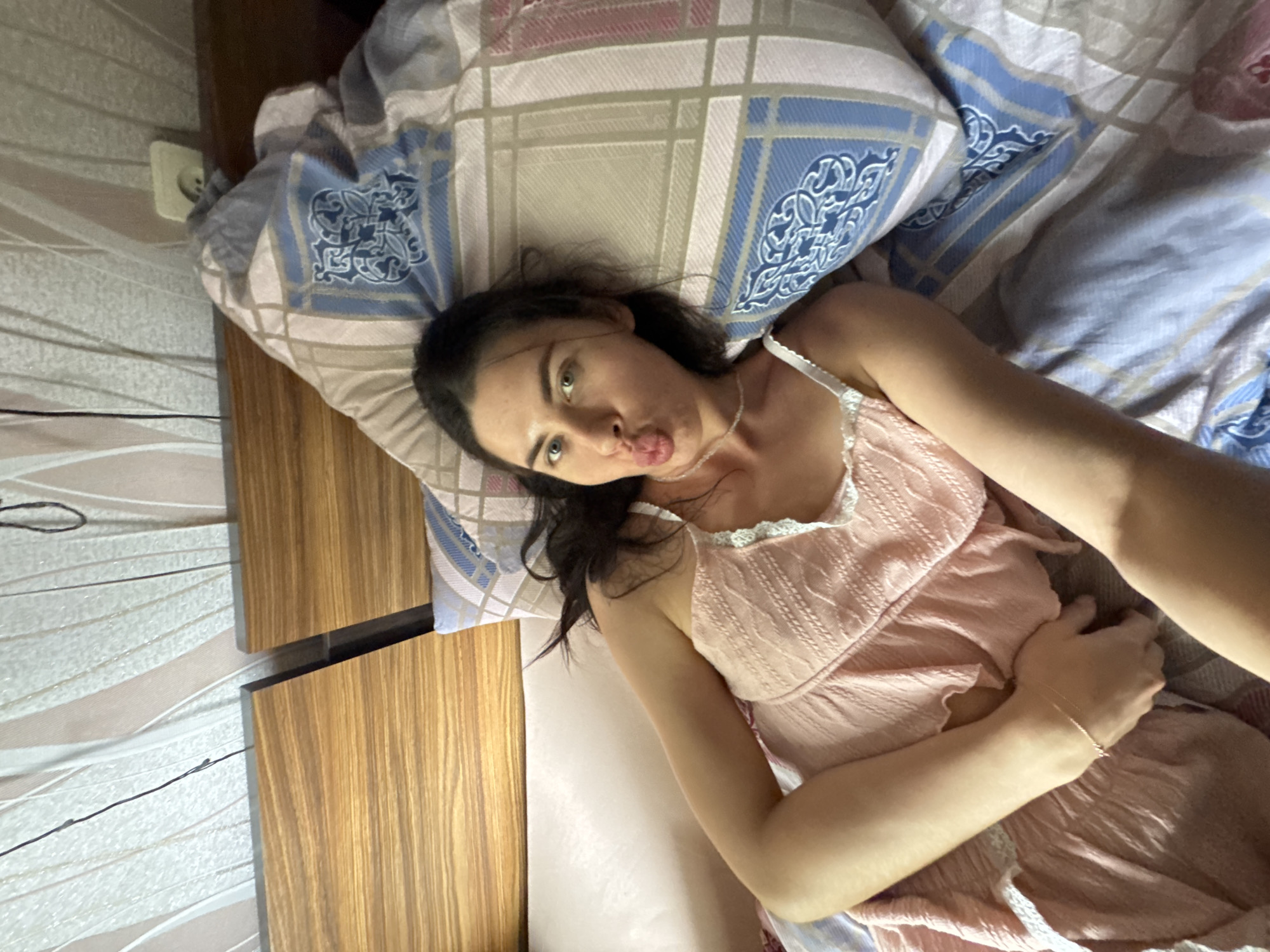 A woman is lying in bed, looking at the camera with her lips puckered. She is wearing a light pink nightgown with lace trim. The bed has wooden headboards and is covered with patterned bedding. The background features a wall with a patterned wallpaper. The scene appears to be an indoor bedroom. No specific location in Chaikovsky, Russia, can be determined from the image.