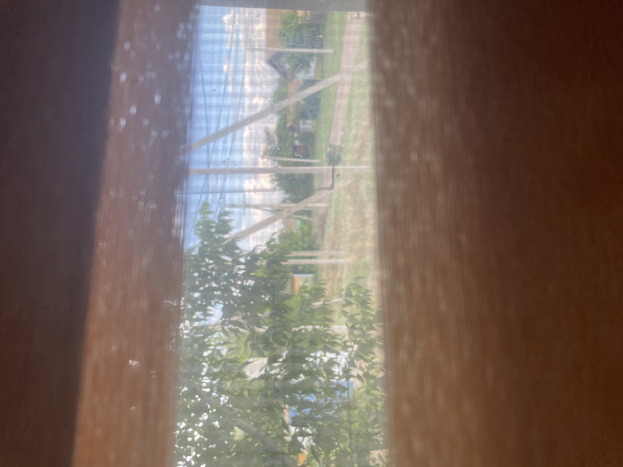 The image shows a view through a window with vertical wooden frames on the left and right sides, creating a tunnel effect. Through the window, a scene unfolds that appears to be an outdoor setting, possibly a rural area in Linyovka, Russia. The perspective is slightly skewed, and the view is somewhat obscured by the texture of the windowpane, which has small water droplets or smudges on it.
In the background, there is a clear blue sky with scattered white clouds, suggesting it is daytime, likely late morning or afternoon given the brightness. The scene beyond the window includes several structures, appearing to be small houses or buildings, amidst lush green trees. There is a path or road winding through the landscape. A greenhouse or a similar structure with white beams and translucent material is visible, stretching across the middle ground.
The foreground is dominated by the blurred textures of the wooden window frames. On the left side, the frame is a dark, rich brown, and it is speckled with small, lighter colored dots, possibly reflections or imperfections on the glass. The right side frame is a lighter, reddish-brown hue and exhibits a similar speckled texture. The foliage of trees with vibrant green leaves is visible in the lower part of the image, suggesting the season is likely summer or late spring. The overall impression is a peaceful, somewhat hazy view of a rural environment. There is no visible text in the image.