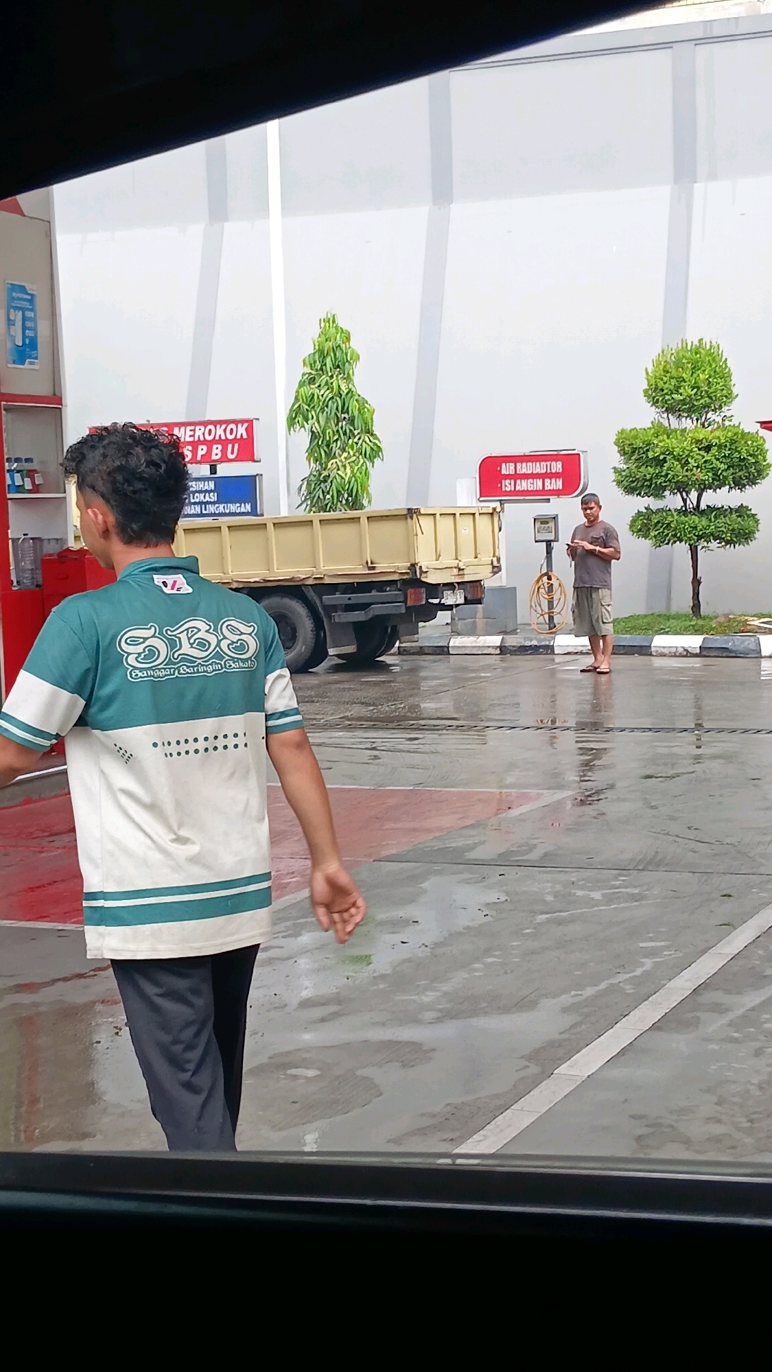 The image depicts a scene at a gas station in Batusangkar, Indonesia. In the foreground, a young man wearing a teal and white polo shirt with the "SBS" logo and dark pants walks away from the camera. Behind him, a yellow truck is parked. To the right of the truck, a man in a gray t-shirt and shorts is standing and looking at his phone. The gas station has white walls, and there are signs indicating "DILARANG MEROKOK" (No Smoking) and "AIR RADIATOR - ISI ANGIN BAN" (Radiator Water - Tire Air Fill). The ground is wet, suggesting it may have recently rained. Several trees are visible in the background. The overall environment appears to be an outdoor commercial area.