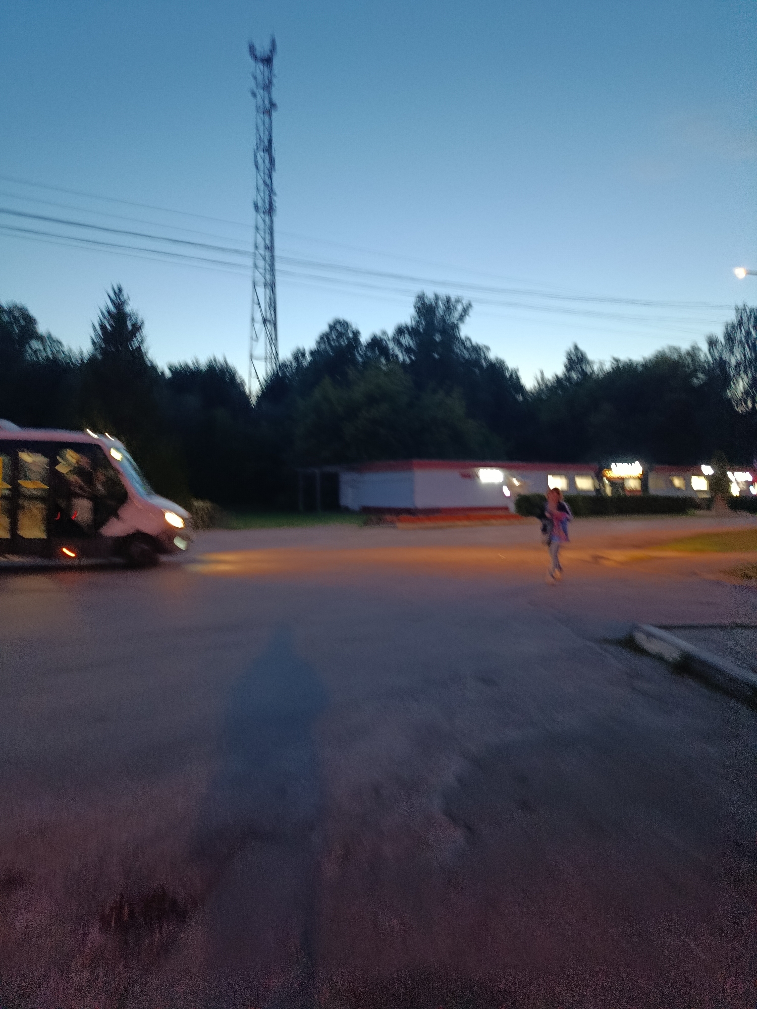 The image depicts a street scene in Verea, Russia, at dusk. A white van is visible on the left, its headlights illuminating the asphalt. Further in the background, a person is walking near a building with illuminated windows. A tall communication tower stands in the distance against the twilight sky. Trees line the background, and overhead power lines are visible. The ground is paved asphalt, with some gravel and debris. The overall impression is of an outdoor, public space transitioning from daylight to night.
