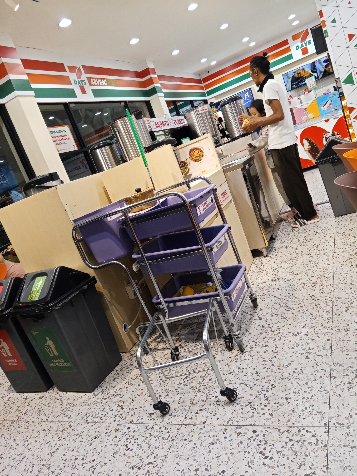 The image depicts the interior of a Seven Eleven convenience store located in Lubuklinggau, Indonesia. The store is brightly lit, with white ceiling tiles and a distinctive red, orange, and green striped logo for "Seven Eleven" visible on the wall. In the foreground, a purple multi-tiered cleaning cart with wheels stands on a terrazzo-patterned floor. To the left of the cart are two large, dark grey trash bins, one with a red label indicating "SAMPAH BOTOL" (bottle trash) and another with a green label for "SAMPAH SISA MAKANAN" (food waste). In the background, a man with long dark hair tied back and a woman are standing near a beverage dispenser. The man is wearing a white t-shirt and dark pants, while the woman is wearing a light-colored top. They appear to be interacting with the dispenser, possibly serving drinks. Several stainless steel beverage dispensers are present, along with other store fixtures and merchandise displays. The overall impression is that of a typical operational convenience store.