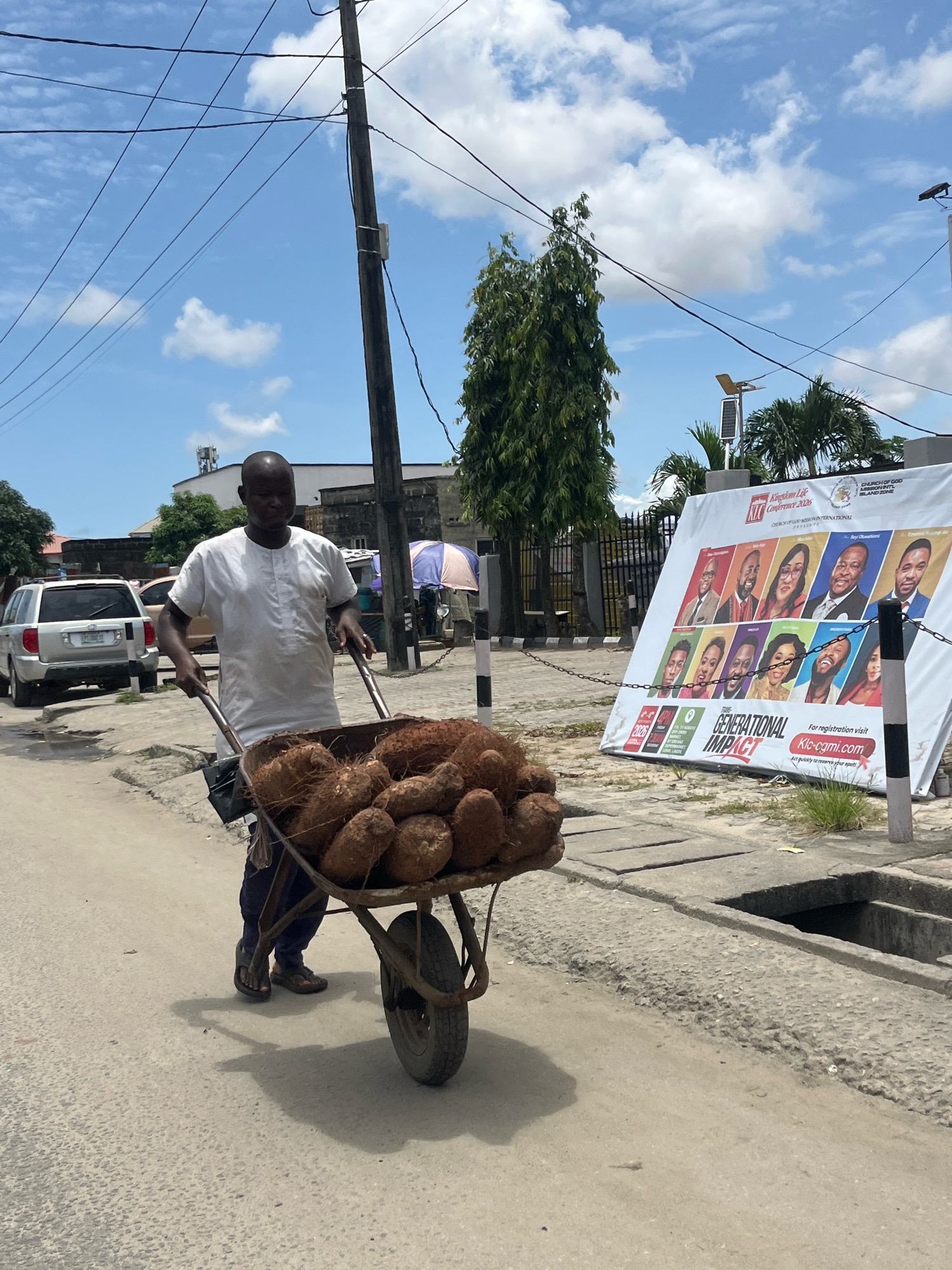 In Ado, Nigeria, a Black man wearing a white kaftan pushes a wheelbarrow filled with yams along a street. The wheelbarrow has a black bag draped over its handles and is loaded with numerous brown, oblong tubers. The man is seen from the waist up, facing slightly away from the camera and looking towards the ground. Behind him, a silver SUV is parked. To the right of the scene, a large white banner for a "Kingdom Life Conference 2026" is displayed, featuring portraits of various individuals and text in English. The environment appears to be an urban street with a power pole, overhead wires, and a drainage ditch visible. The sky is blue with scattered clouds.