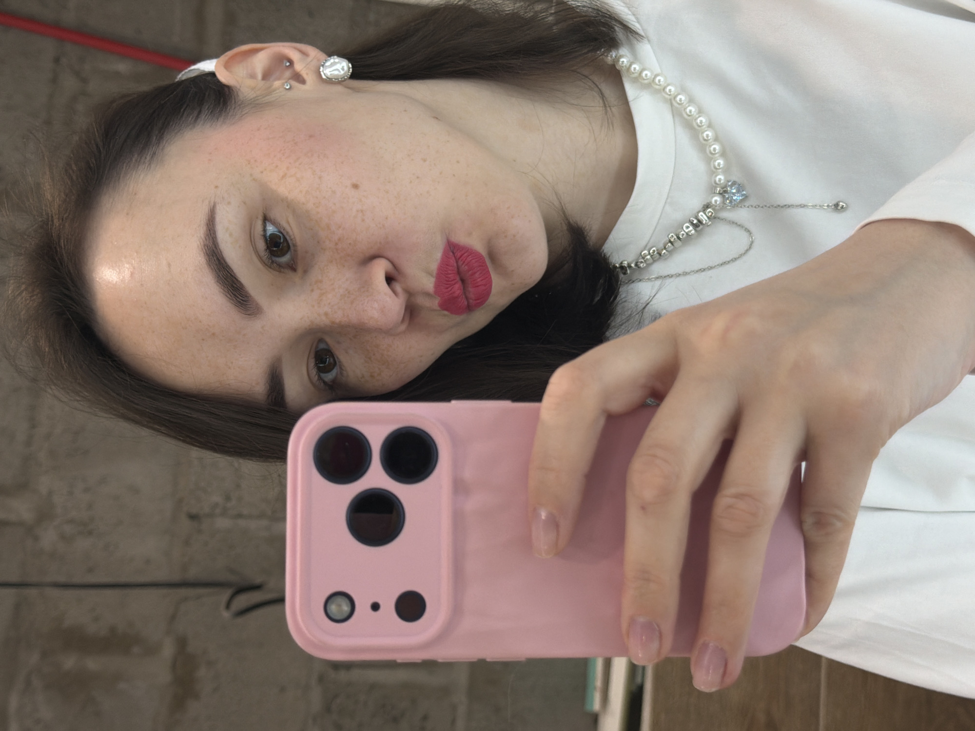 A woman with brown hair is taking a selfie with a pink smartphone. She is wearing a white shirt and a pearl necklace with a silver pendant. Her lips are red, and she has freckles on her face. She has several piercings in her left ear. The background is blurry, but it appears to be a room with neutral-colored walls. There is a red object in the upper left corner of the frame. The location is unknown, and there is no information in the image to indicate it is Chaikovsky, Russia.