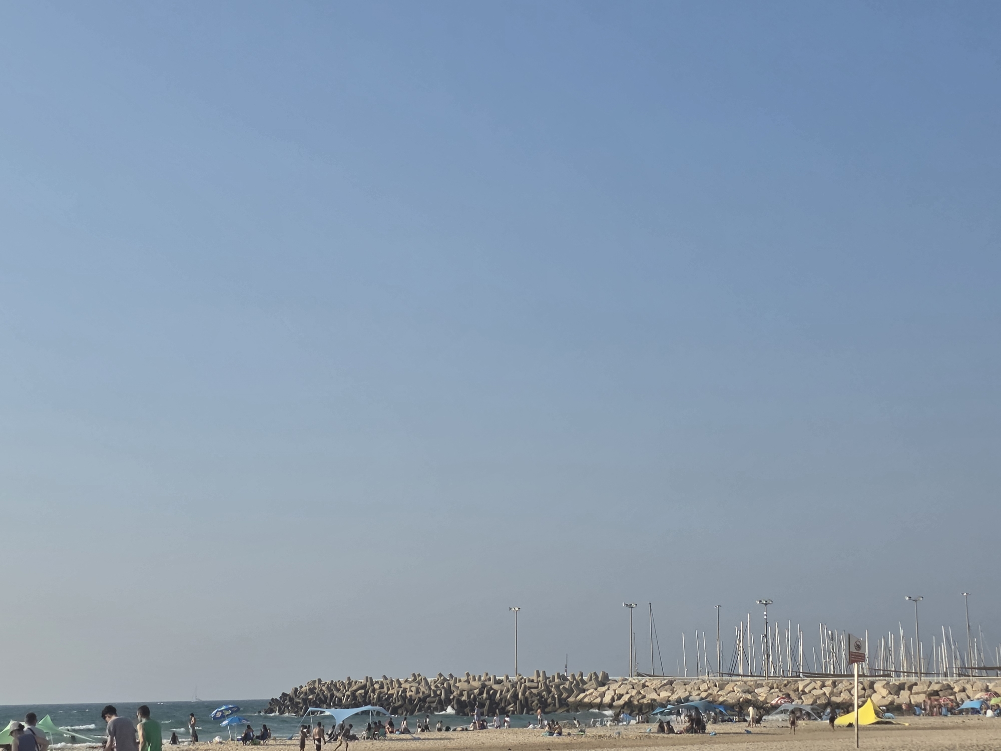The image captures a sunny day at a beach in Ashkelon, Israel. The foreground shows a sandy beach with numerous people relaxing under umbrellas and sunbathing. Some individuals are seen walking along the shore. In the mid-ground, a man-made breakwater composed of large concrete blocks separates the beach from a body of water. Beyond the breakwater, numerous sailboat masts are visible, indicating a marina or harbor. Two lampposts stand on top of the breakwater, and a yellow flag is planted on the sand near the right edge of the frame. The sky is clear and blue, with no clouds present.