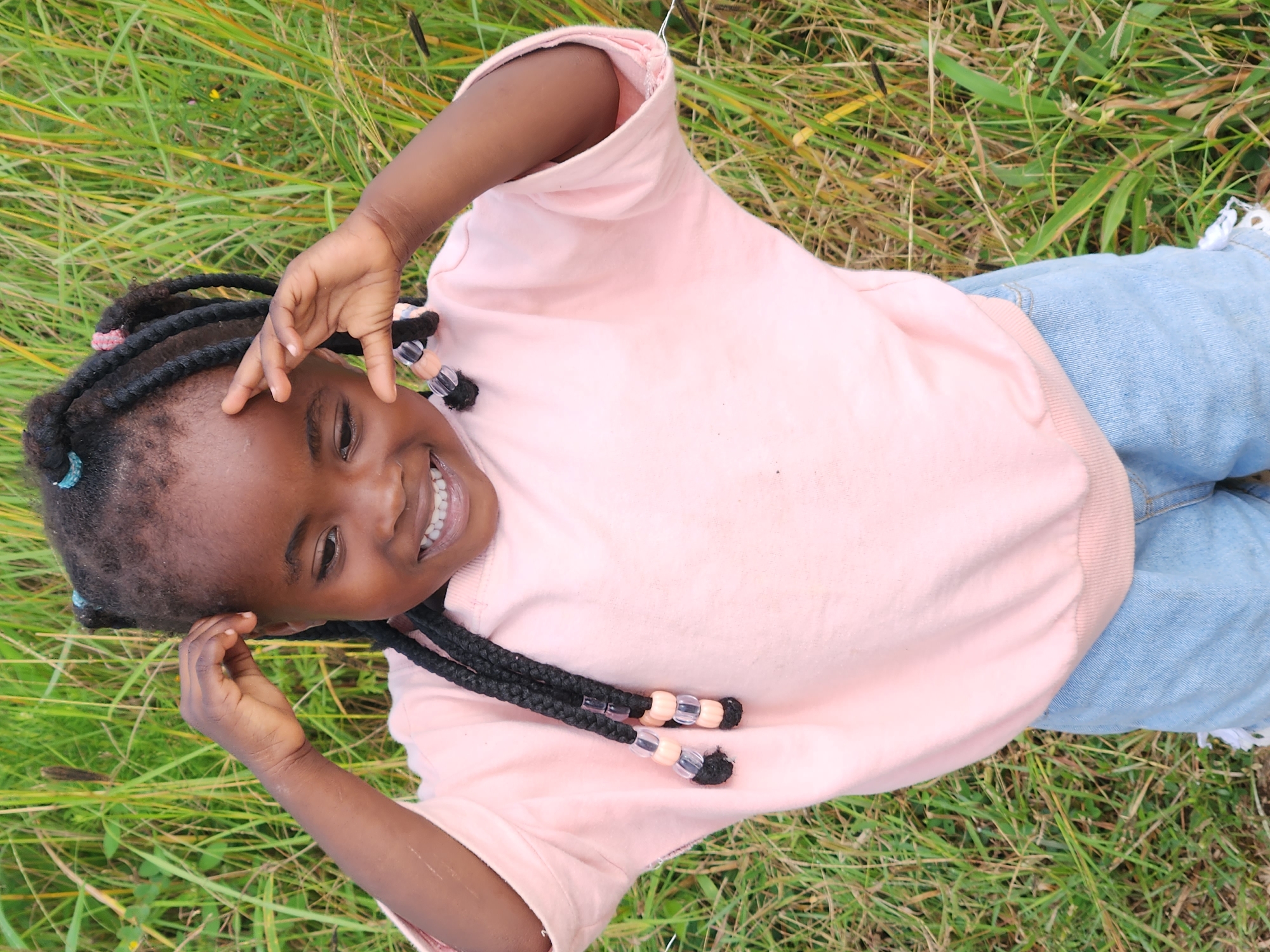 A young Black girl with braided hair adorned with beads stands in a field of tall, green grass. She is smiling broadly and has her hands raised to her temples. She is wearing a light pink sweatshirt and denim shorts. The background is blurred, but it appears to be a natural outdoor setting. The lighting suggests it is daytime, likely with natural sunlight.