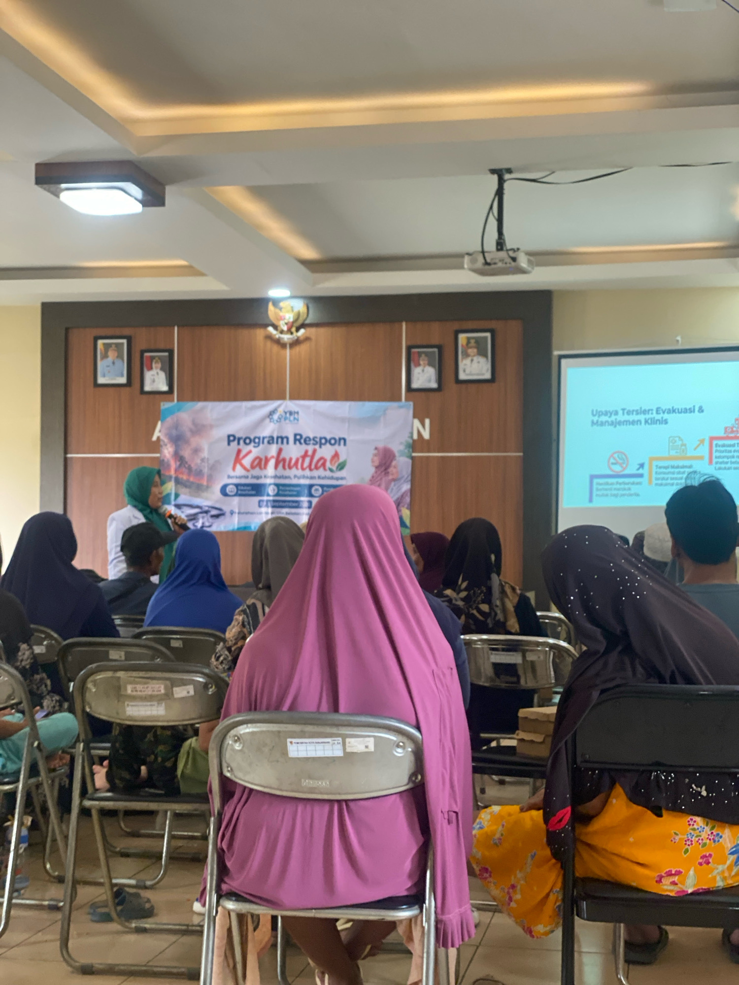 The image depicts an indoor event in Banjarbaru, Indonesia, likely at a government or community building, given the presence of official portraits on the wall and a projector screen. A group of people, mostly women wearing hijabs, are seated, facing a speaker who is standing near a banner. The banner, in Indonesian, reads "Program Respon Karhutla," suggesting a program related to forest and land fires. The speaker, a woman in a green hijab, is holding a microphone and addressing the audience. A projector screen displays text and graphics, indicating a presentation is underway. The room appears to be set up for a meeting or seminar, with chairs arranged in rows and a podium or small table near the speaker. The lighting suggests it is daytime, but the ambient light from the ceiling fixtures is also visible. The overall atmosphere seems formal, with an educational or informational purpose.