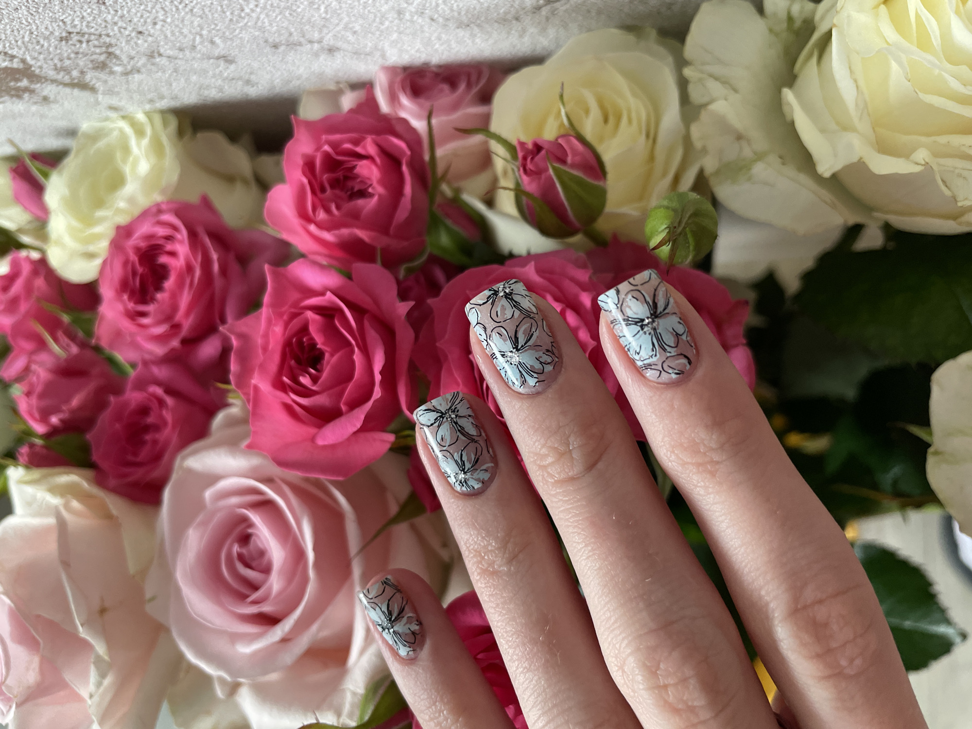 A close-up shot features a person's hand with elaborately manicured nails, positioned gracefully among a vibrant bouquet of roses. The hand, seen from the wrist upwards with fingers extended, showcases nails adorned with a delicate floral design. Each nail displays outlines of flowers in dark lines, filled with a pale blue or greyish-white color, with some flowers featuring small, sparkling rhinestones or gems at their centers.
The background is dominated by a lush arrangement of roses in various stages of bloom and colors. Bright fuchsia pink roses are prominent, intermingled with lighter pink and creamy white roses. Several rosebuds, some still tightly closed, add to the natural beauty of the floral display. The rich greens of rose leaves are visible peeking through the blossoms. At the very top of the frame, a textured, light-colored surface, possibly a wall or decorative box, provides a soft, neutral backdrop.
The scene suggests an appreciation for beauty and detail, possibly a personal aesthetic shot, showcasing the nail art in a natural, elegant setting. The lighting appears soft and natural, indicating daytime. No visible text is present in the image.