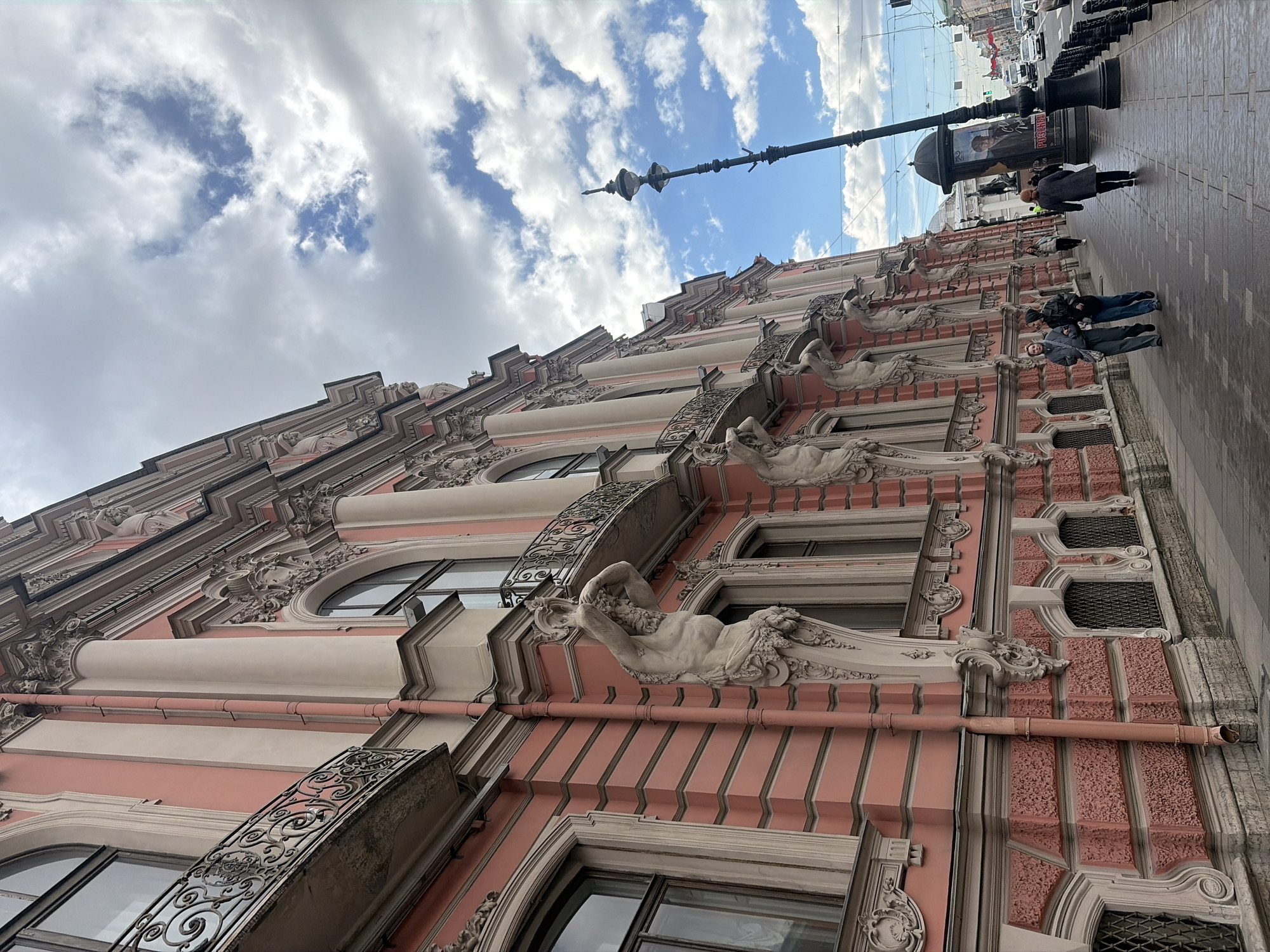 An outdoor, low-angle shot captures a vibrant street scene in Saint Petersburg, Russia, dominated by the elaborate facade of a historic building under a partly cloudy sky. The multi-story building features a distinctive pinkish-red main color, intricately adorned with white and cream architectural details. These details include ornate moldings, decorative balustrades, and a series of sculpted reclining figures, reminiscent of Atlantes or Caryatids, supporting projections above the windows. The facade also displays arched and rectangular windows, some with decorative wrought-iron balconies. A reddish-orange drainpipe runs across the building's surface.
To the right of the building, a paved sidewalk stretches into the background. Several pedestrians, dressed in coats, walk along the sidewalk. Prominent street furniture includes a tall, black ornate lamppost and a cylindrical street advertisement kiosk. The kiosk displays various posters, with the Cyrillic word "PITER" clearly visible on one of them. In the distance, a street with vehicles can be discerned. The sky above presents a cheerful mix of bright blue patches and fluffy white clouds, indicating clear daytime weather.
