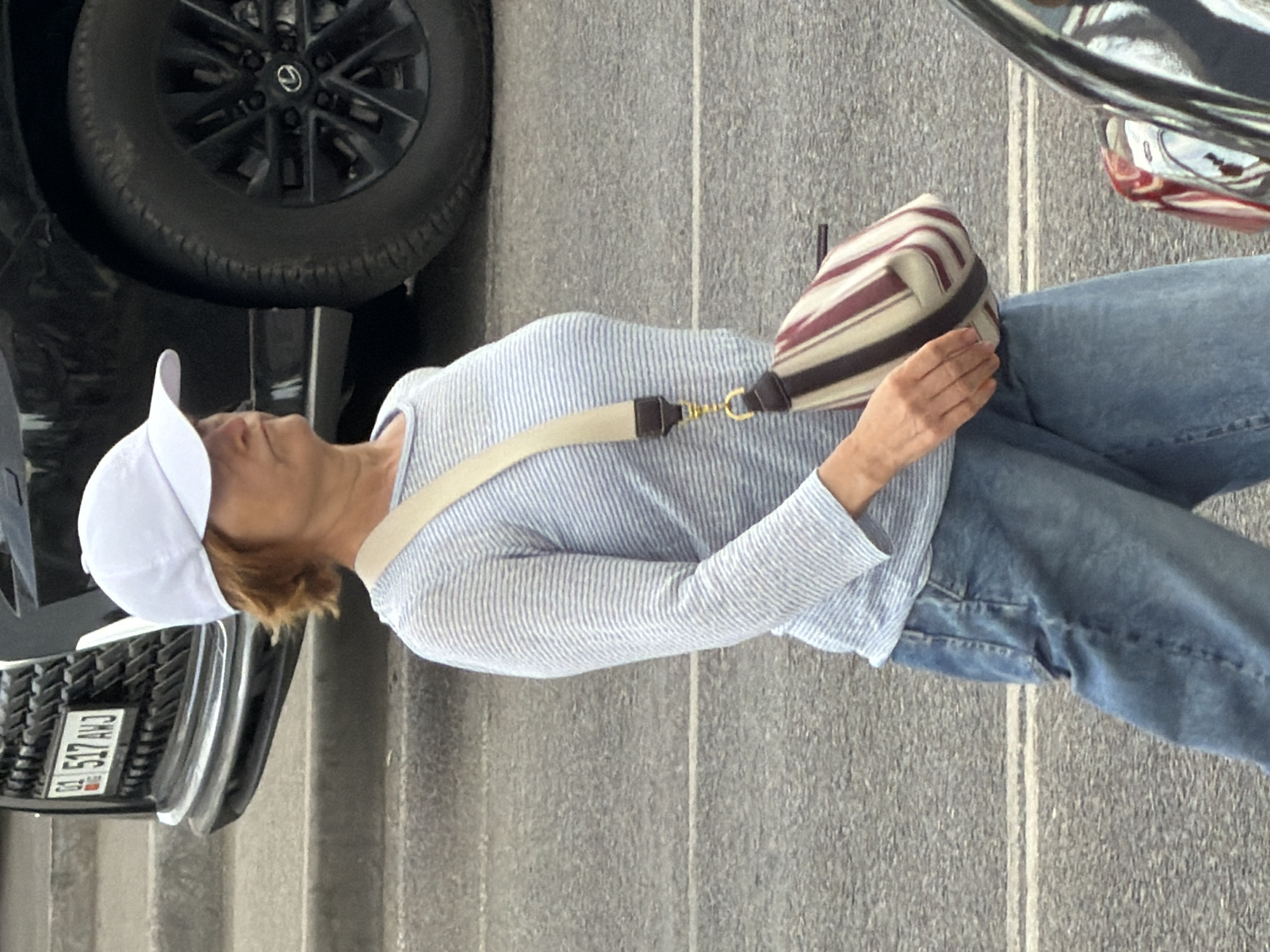 A woman with shoulder-length, reddish-brown hair wears a white baseball cap, a light blue and white striped long-sleeved shirt, and loose-fitting blue jeans. She carries a crossbody bag with a light-colored strap and a bag featuring vertical stripes of burgundy, off-white, and dark brown. The woman is walking on a grey paved surface, likely a street or sidewalk, with a white line painted on the ground. Behind her is the front right quarter of a black SUV with a silver license plate frame and a black grille. Part of the SUV's tire and a portion of its bumper are visible. A small red and chrome object, possibly part of another vehicle, is visible in the lower right corner of the frame. The scene appears to be outdoors in daylight.