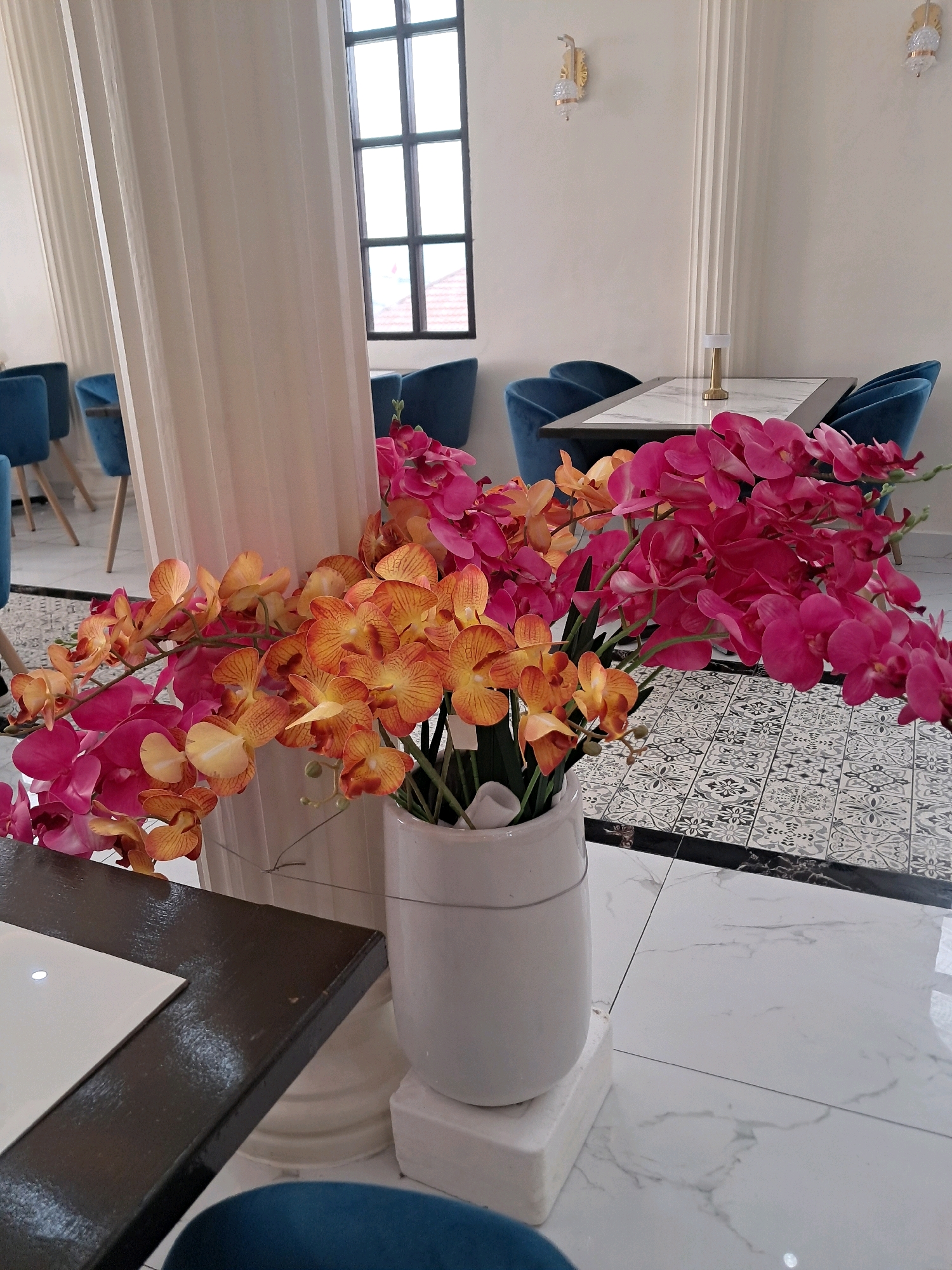 The image shows a close-up view of a vase of artificial orchids, with pink and orange blooms, placed on a white surface in the foreground. Behind the vase, a tall, fluted white pillar is visible, obscuring a portion of the background. In the background, there are several blue chairs arranged around tables, and a window with a grid pattern. The floor is tiled with a black and white geometric pattern, with white marble-like tiles elsewhere. The scene appears to be set in an indoor dining area or cafe. There are no people or animals visible in the image. The lighting suggests an indoor setting with natural light coming from the window. The overall impression is one of a decorative display in a public space.
