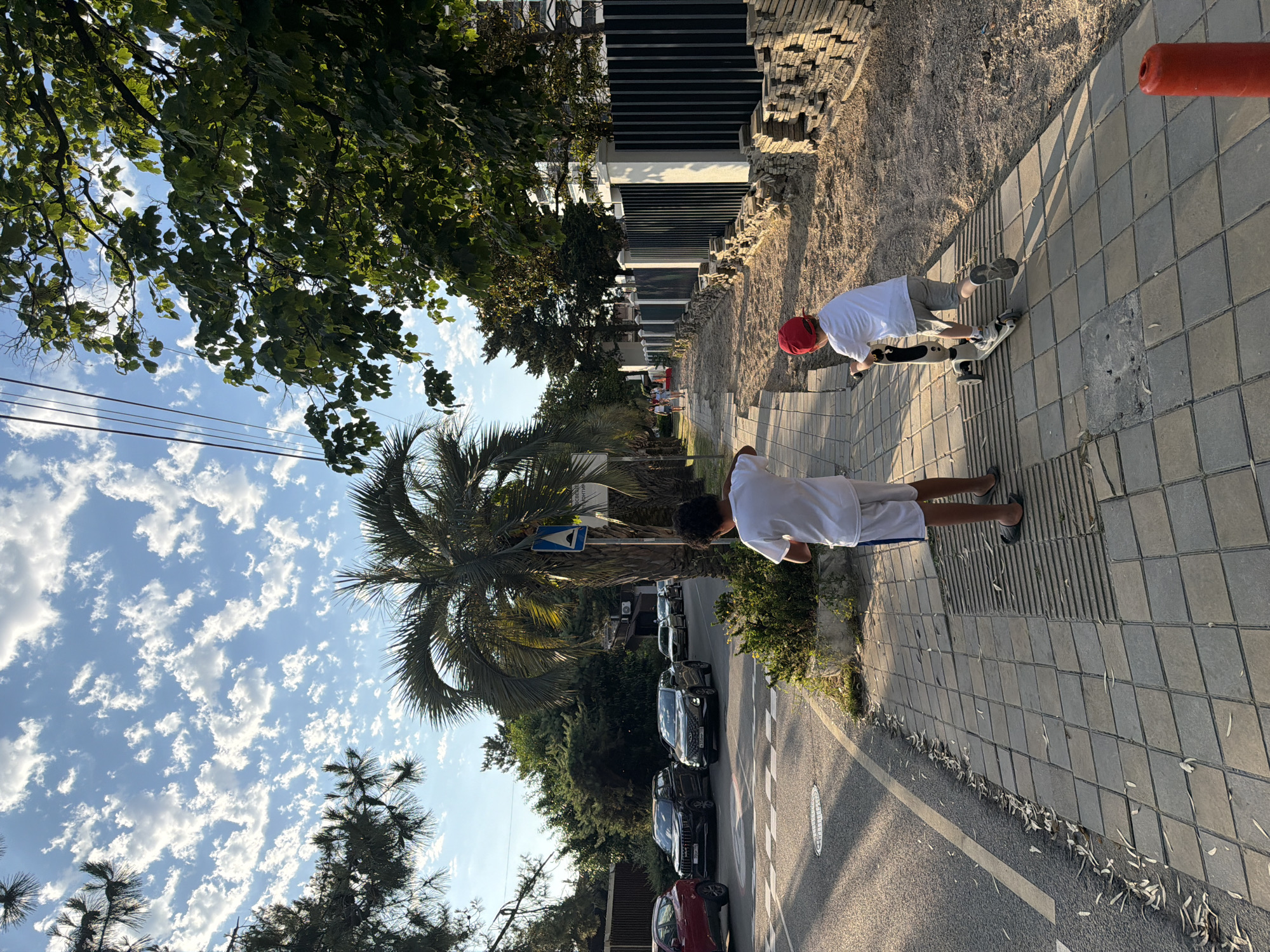 The image depicts a street scene in Sochi, Russia, on a sunny day with scattered white clouds. In the foreground, two boys are present. One boy, facing away from the camera and wearing a white t-shirt and white shorts with blue trim, stands on a tiled sidewalk. He is wearing dark flip-flops. Behind him, a second boy, also facing away from the camera, is riding a scooter. He is wearing a white t-shirt, grey shorts, and a red baseball cap. The scooter is white and grey. The sidewalk is made of large square grey tiles.
To the left of the sidewalk is a road with cars parked and driving. Several vehicles are visible, including a red SUV, a black SUV, and a silver sedan. The road has yellow and white lane markings. To the right of the sidewalk, there is a sandy area with stacks of stone tiles, suggesting ongoing construction or landscaping. A black metal fence with white concrete posts runs along the sandy area.
Palm trees and other green foliage are prominent throughout the scene, with tall trees and lush bushes lining the street and the properties beyond. In the background, buildings are visible, including a white building with dark windows. Overhead power lines stretch across the frame. The lighting indicates it is late afternoon, with long shadows cast by the trees and people.