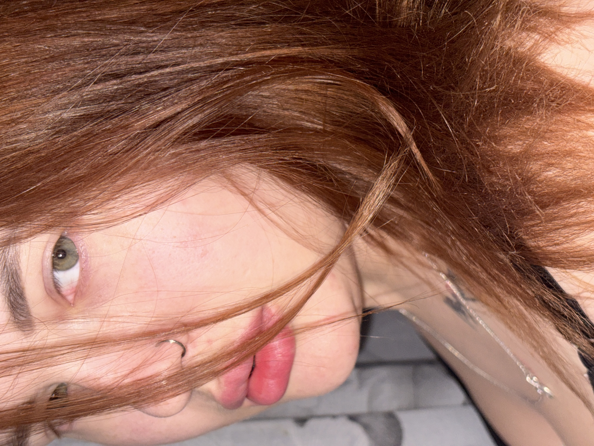 A close-up, tilted view of a person's face is presented. The individual has light skin, reddish-brown hair, and green eyes. Part of the face is obscured by strands of hair. A nose ring is visible. A silver necklace can be seen around the neck. The background appears to be a textured, possibly marble-like surface. The location and specific actions or interactions in the scene are not discernible. The city of Novomargaritovo, Russia is not featured.