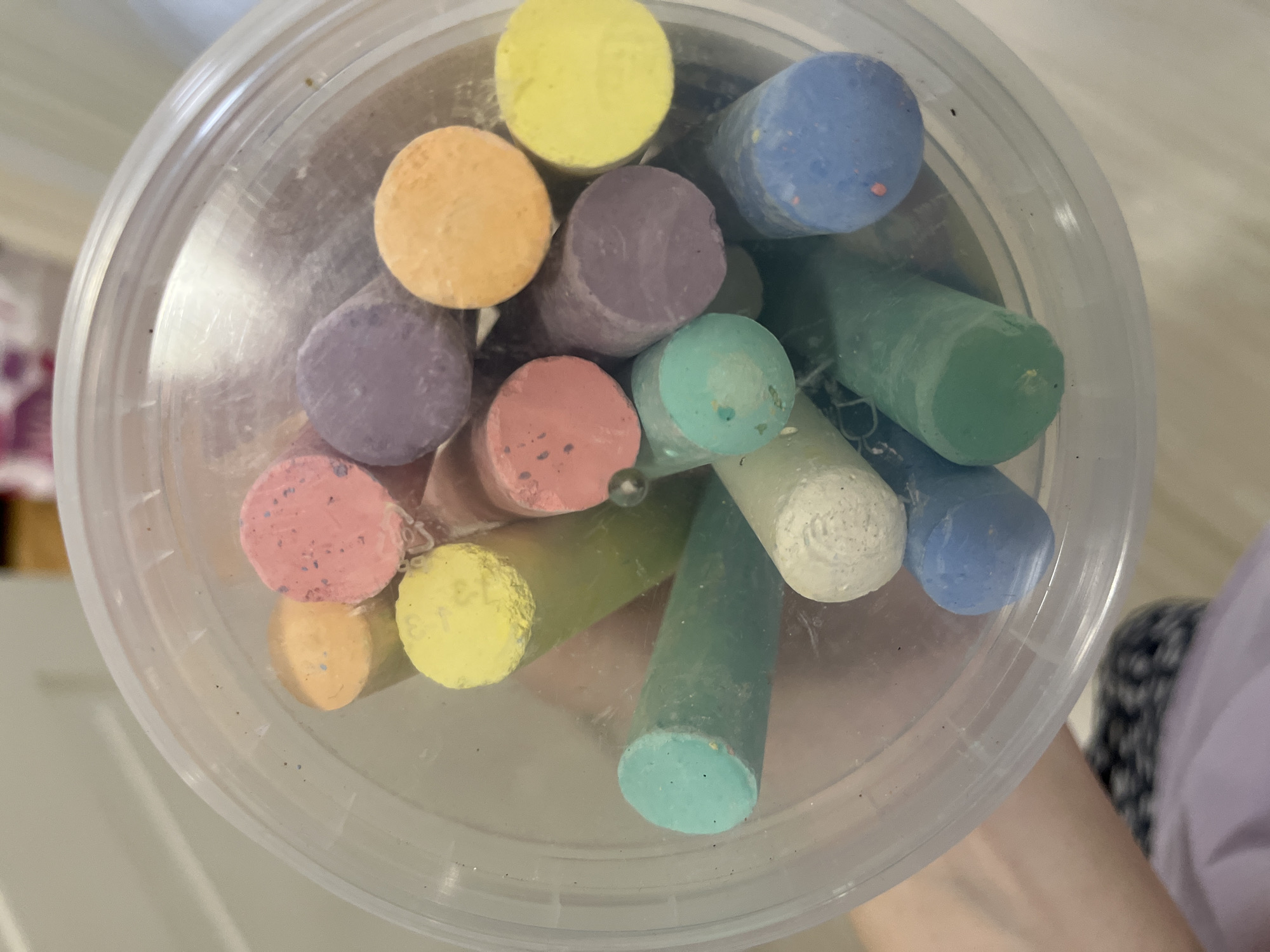 The image shows a clear plastic container filled with colorful chalk sticks. The chalk colors include yellow, orange, purple, pink, green, blue, and white. The container appears to be round with a slight ridge near the top. There's a hint of a possible sleeve or shirt visible in the lower right corner. The lighting is natural and even. There is no way to determine from this image if it was taken in Chaikovsky, Russia.