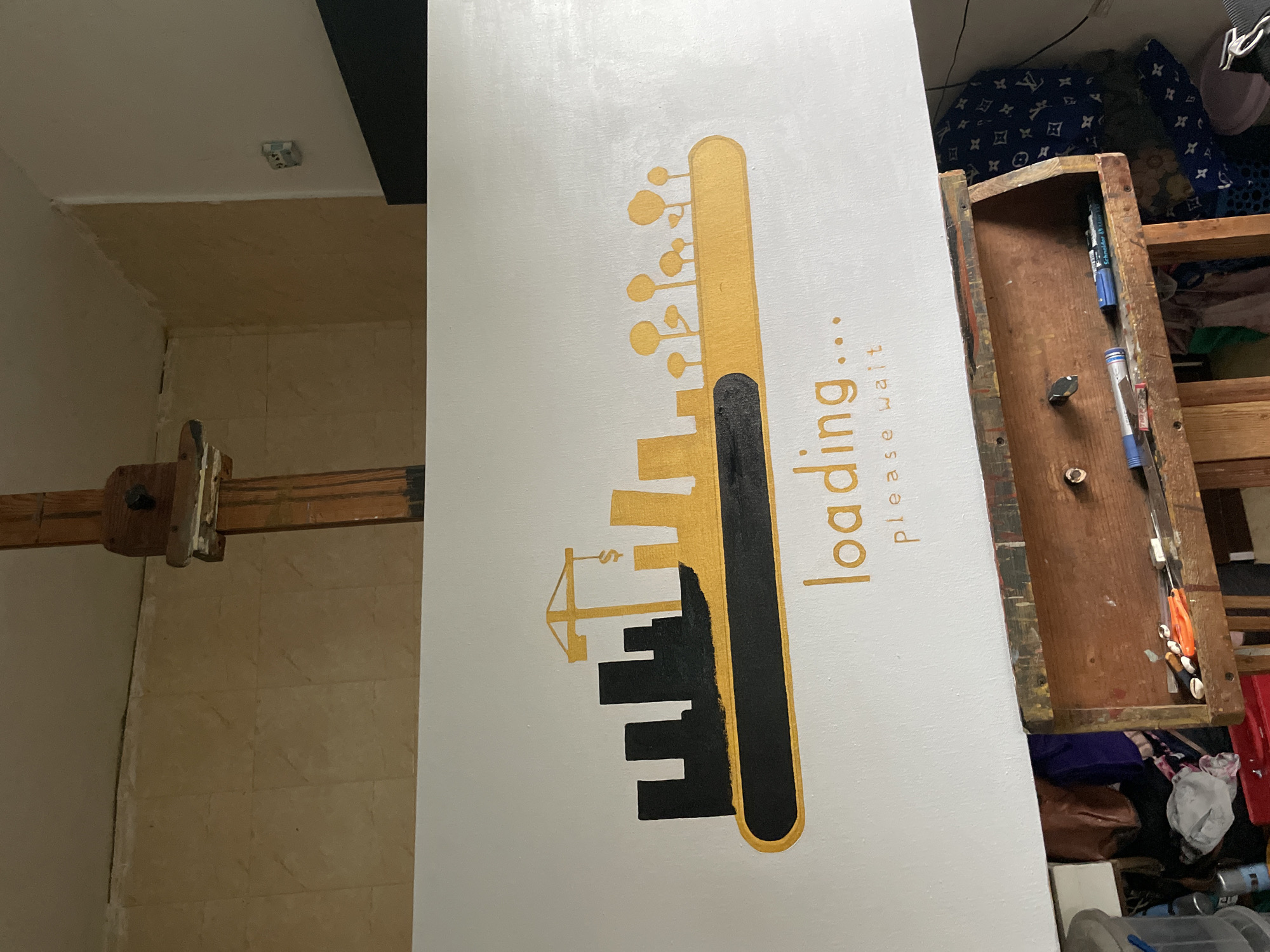 The image shows a partially visible room with tiled flooring. A wooden beam with metal fixtures is mounted on a wall. A white canvas featuring a graphic in gold and black depicting a city skyline in the shape of a key with "loading..." and "please wait" text is propped against an open, cluttered wooden drawer or shelf. The drawer contains tools and other miscellaneous items. In the background, various items are stacked on shelves, including bags, clothes, and other storage containers. The location of Ado, Nigeria is not evident from the visual information provided.