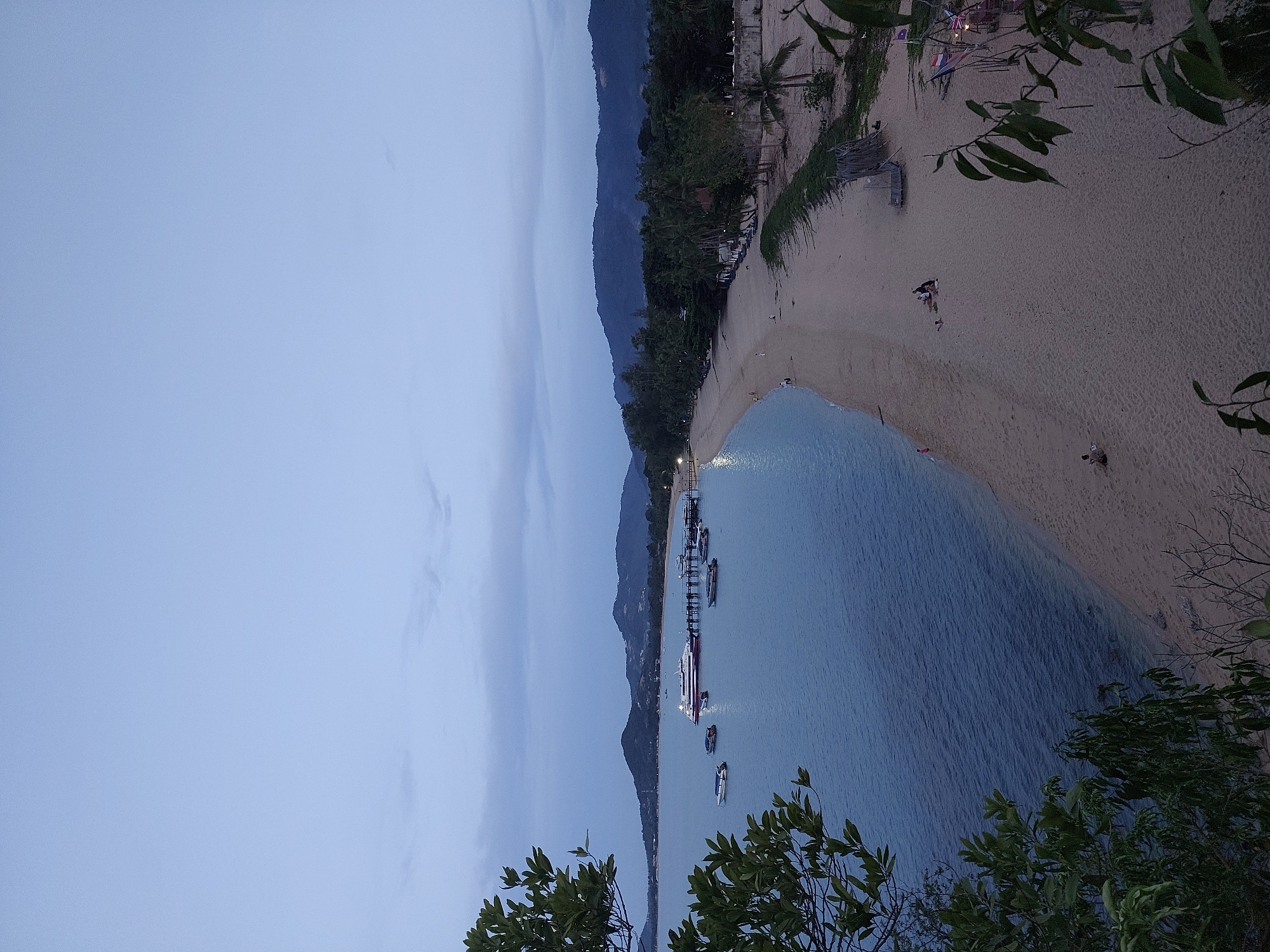 The image depicts a scenic view of a beach and ocean at dusk. The scene is captured from a slightly elevated perspective, with lush green foliage framing the bottom and right edges of the frame. The sky is a pale blue, indicating the time of day is late evening, shortly after sunset.
The beach, a soft expanse of sand, curves gently along the coastline. Several people are visible on the sand, scattered in small groups, suggesting a leisurely and relaxed atmosphere. Some appear to be walking, while others are seated or engaged in quiet conversation.
The ocean, a calm and inviting expanse of blue water, laps at the shore. A pier extends out into the water, where several boats are moored. One larger boat, possibly a ferry or a smaller cruise ship, is prominently docked, its lights beginning to illuminate the darkening water. Smaller boats, likely speedboats or personal watercraft, are also visible.
In the background, a mountain range rises, covered in dense vegetation. The outlines of the mountains are softened by the twilight. Small structures, possibly huts or resort buildings, are nestled along the edge of the beach, hinting at the presence of tourist accommodations. Small flags, including what appears to be a Union Jack, are visible on the beach, suggesting a connection to international visitors.
The overall mood of the image is peaceful and serene, capturing the tranquil beauty of a tropical beach as day transitions into night. There is no visible text in the image.
