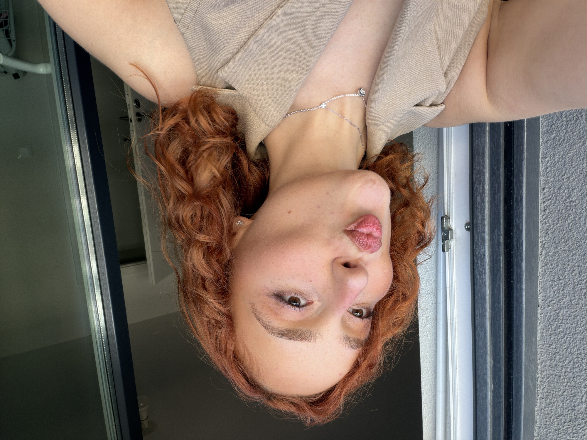 A young Caucasian woman with vibrant red, curly hair is captured in a close-up, selfie-style photograph. She is wearing a beige sleeveless blazer and a delicate silver necklace with a heart-shaped pendant. Her lips are pursed in a "duck face" pose, and she is wearing subtle makeup, including eyeshadow and blush. She has freckles across her nose and cheeks.
The background suggests an outdoor setting, possibly a balcony, with a gray textured wall and a window frame visible on the left. On the right, a dark, reflective glass door or window is present, showing a blurred interior with what appears to be a white railing or crib in the distance. The lighting indicates it is daytime.
While the image does not provide specific clues to pinpoint the exact location within Strasbourg, France, the architectural elements of the building and the overall ambiance are consistent with residential buildings found in the city.