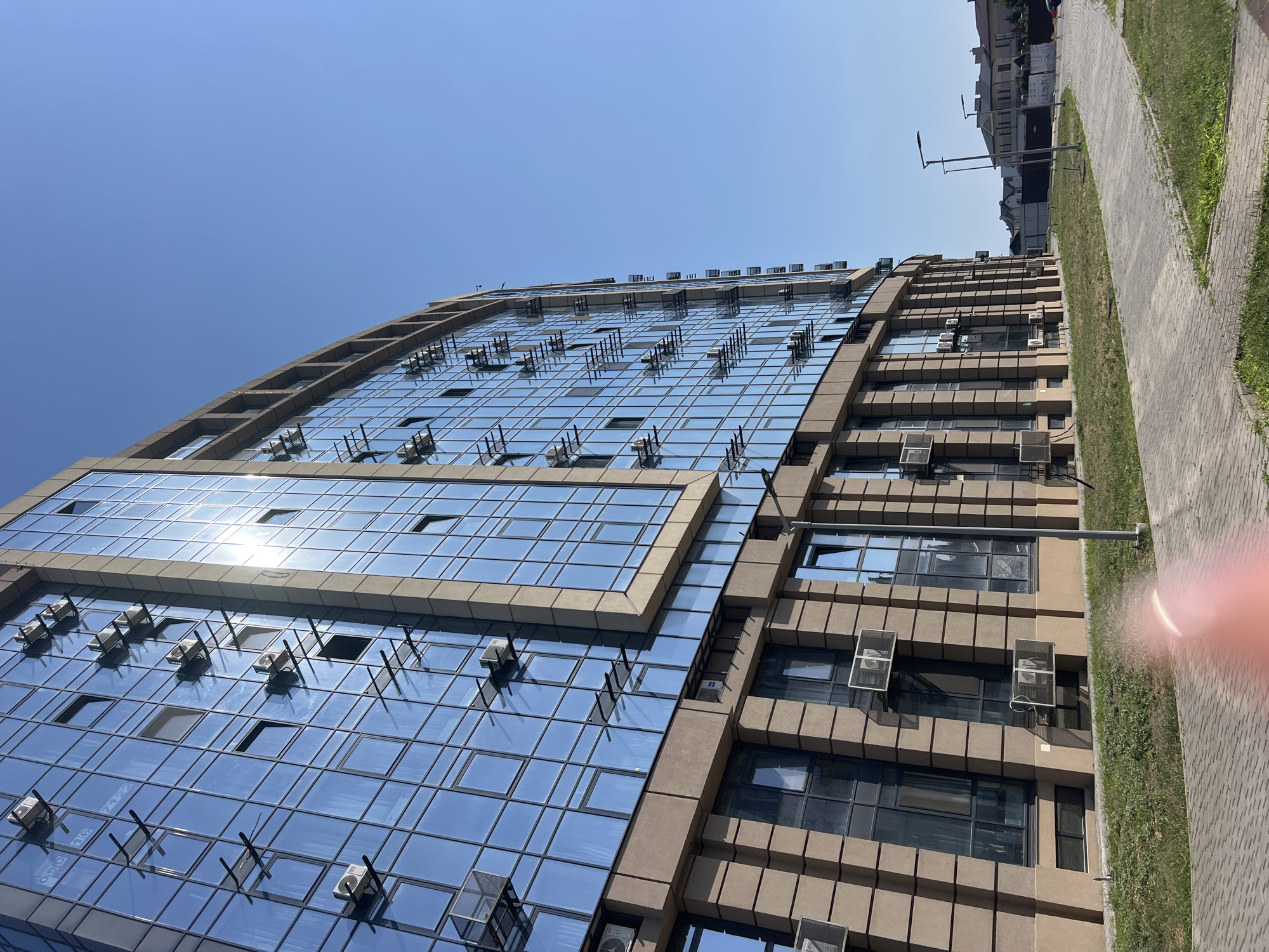 This is a low-angle, eye-level, full shot of a modern, multi-story building with a blue, reflective glass facade and beige stone accents, located in Fontanka, Ukraine. The building is surrounded by a paved walkway and a grassy area. The sky is clear and blue. Numerous air conditioning units are mounted on the exterior of the building. In the background, residential buildings and streetlights are visible. There is no human presence in the image.