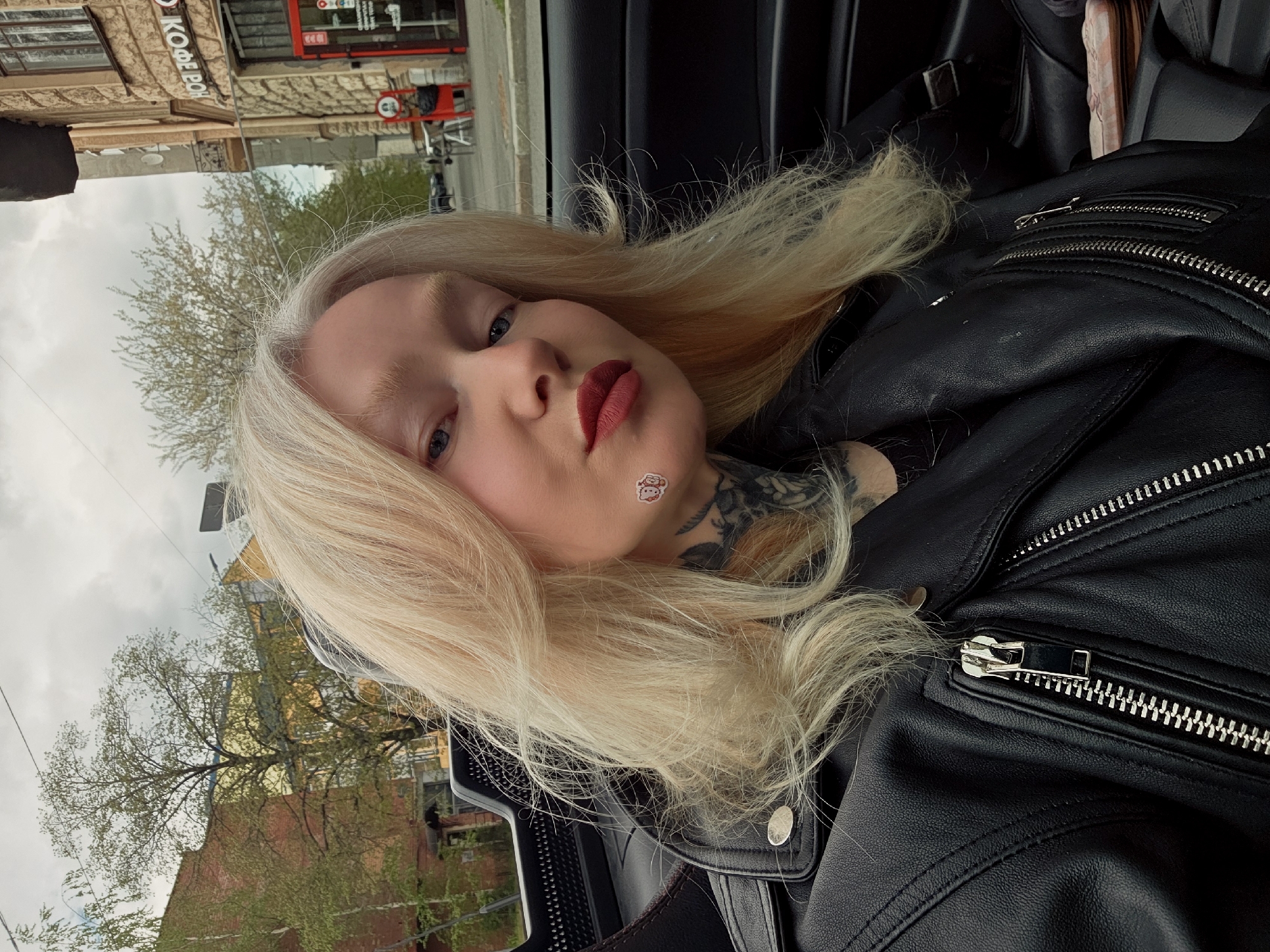 A medium shot shows a white woman with long blond hair and light skin. She has a sticker on her neck. She is wearing a black leather jacket. The photo appears to have been taken inside a car. A building is visible in the background with lettering in Russian script, specifically "КОФЕВОИ...", suggesting a cafe with coffee. Also visible in the background is foliage, and it's possible to infer a street scene. The scene suggests the city of Saint Petersburg, Russia.