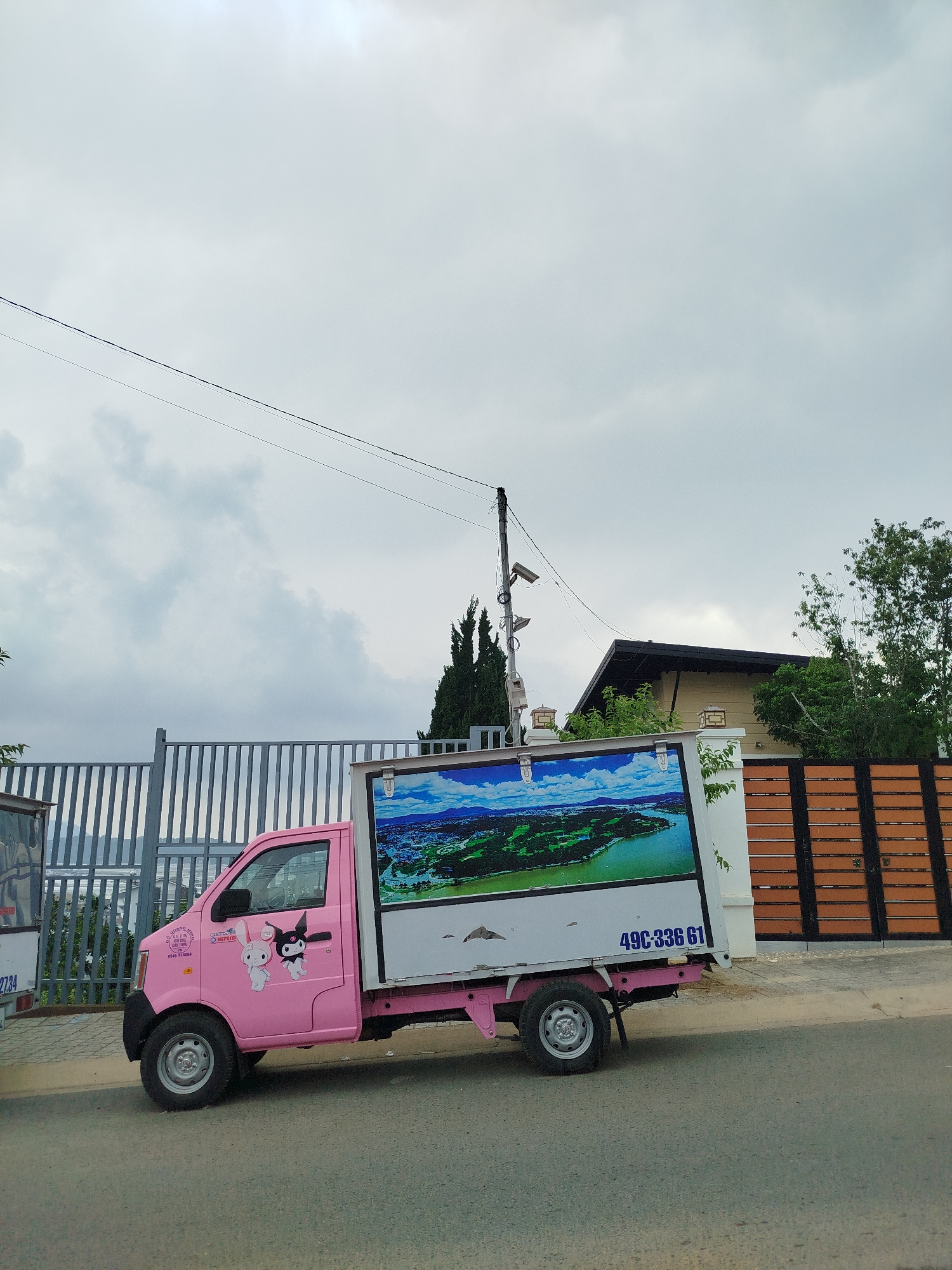 A pink mini truck with cartoon characters and a large advertisement on its side is parked on a paved road in Lâm Viên - Đà Lạt, Vietnam. The truck features an image of a landscape with water on its side. The number "49C-336 61" is printed on the back of the truck. The road is bordered by a tall metal fence on one side and a shorter wall with a brown wooden gate on the other. A surveillance camera pole stands behind the wall. The sky is overcast.