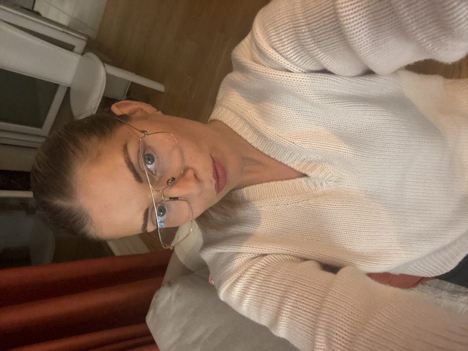 A woman in glasses and a white sweater poses for a selfie. Her hair is pulled back, and she looks directly at the camera with a slight tilt of her head. The background includes a glimpse of a window and a red curtain. The lighting appears soft and diffused. The sweater's texture is visible, and the gold frames of the glasses stand out against her face.