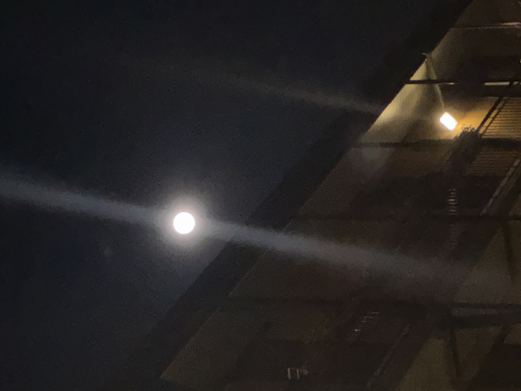 A nighttime shot in what appears to be Milan, Italy, shows the moon shining brightly in the night sky, with its light reflecting off the surrounding environment. The moon has a light-ray effect. Architectural details are visible in the right side of the frame, possibly part of a building's structure. One artificial light source can be seen from a nearby building. The overall mood is quiet and atmospheric.
