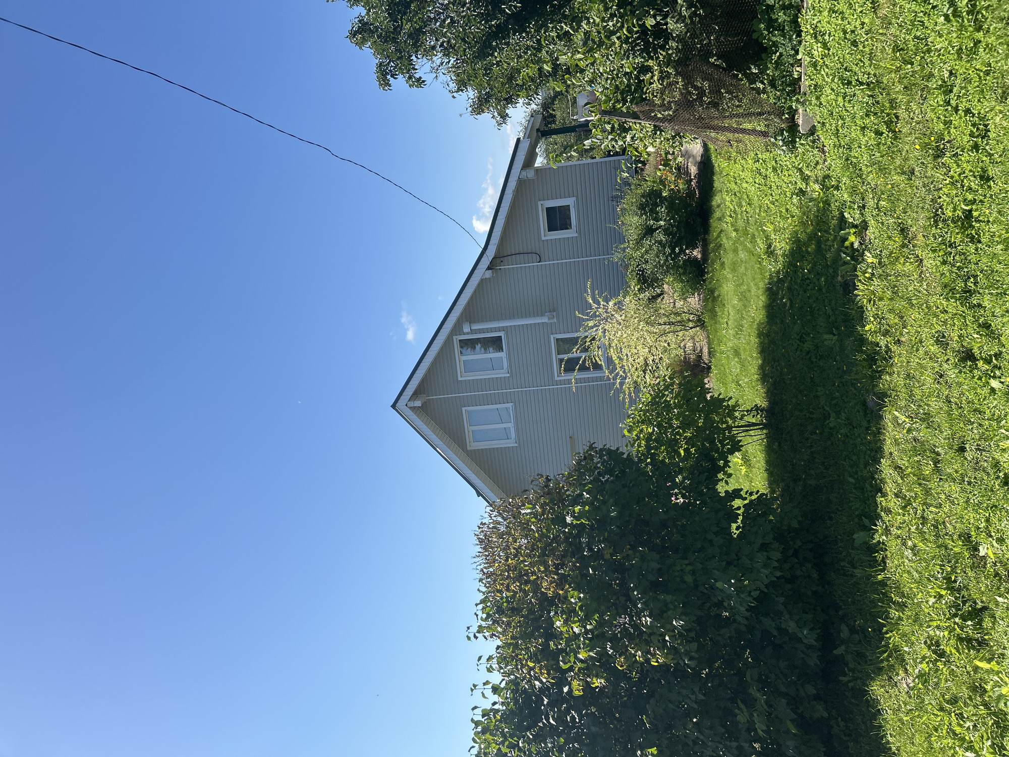 The image captures a two-story light gray house with a dark roof, set against a clear blue sky. The house features several white-framed windows, two on the upper floor and three visible on the lower floor. A white pipe runs vertically down the side of the house. The surrounding environment is lush and green, with a well-maintained lawn in the foreground and various bushes and trees bordering the property. In the background, a dark wire stretches across the sky. There are no people or animals visible in the frame. The scene suggests a peaceful, rural or suburban setting, possibly in the city of Kalitsino, Russia, given the context.