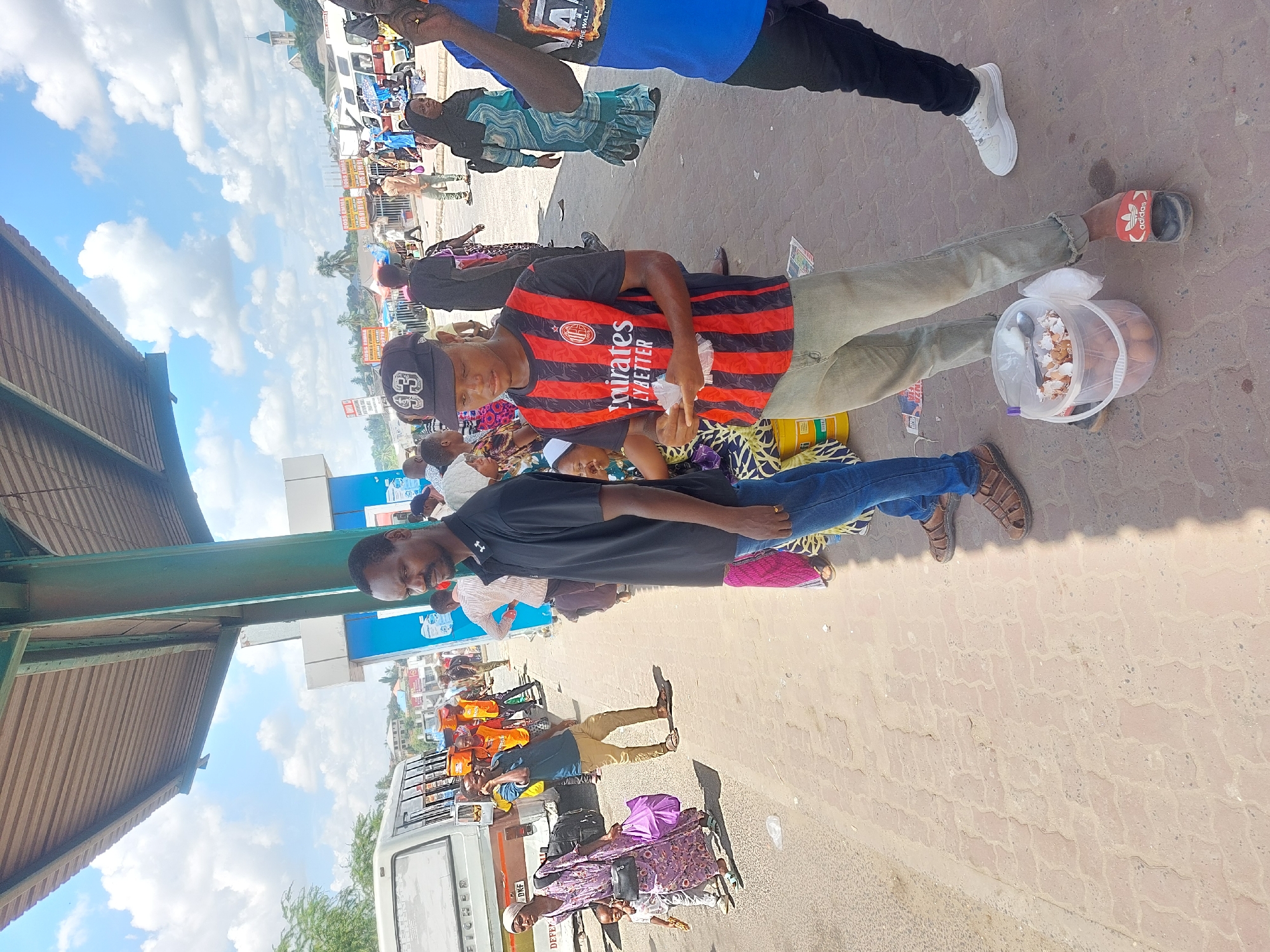The image depicts an outdoor scene at what appears to be a busy transportation hub in Dar es-Salaam, Tanzania. Several people are visible, some standing and some sitting on the ground, with various belongings surrounding them.
A man wearing a black and red striped jersey is seen holding something in his hand, possibly food or money. Beside him, another man is wearing a plain black shirt and appears to be looking directly at the camera. A plastic bucket full of eggshells and eggs sits on the ground.
A white van is parked in the background, with other people gathered nearby. The environment suggests a bustling marketplace or transport station. Visible in the distance are buildings and structures characteristic of an urban setting. The overall ambiance of the scene indicates a typical day in a busy urban environment in Dar es-Salaam.