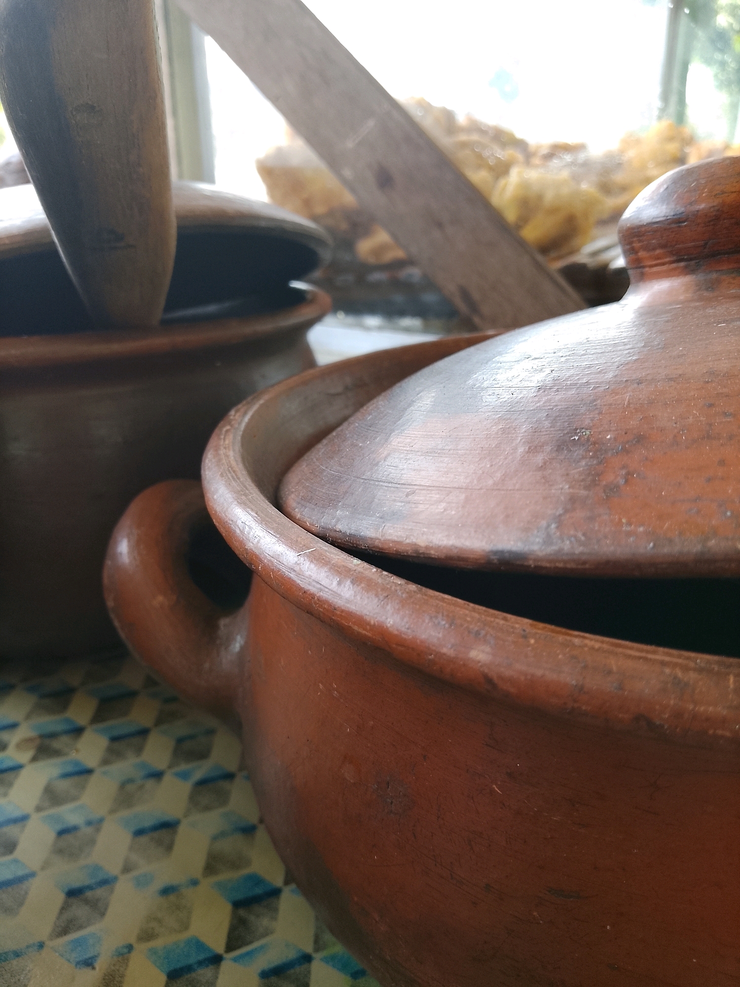 This is a close-up, angled shot of two earthenware pots with lids, placed on a patterned surface. The pot in the foreground is a reddish-brown color and has a handle. Its lid is slightly ajar, revealing a dark interior. A wooden utensil is visible resting on the lid. Behind it, another similar pot is visible, also with a lid, appearing darker in color. The background is blurred, showing a window with light streaming through, and hints of greenery outside, suggesting an indoor setting near a window, possibly in a kitchen or dining area in Banaran, Indonesia.