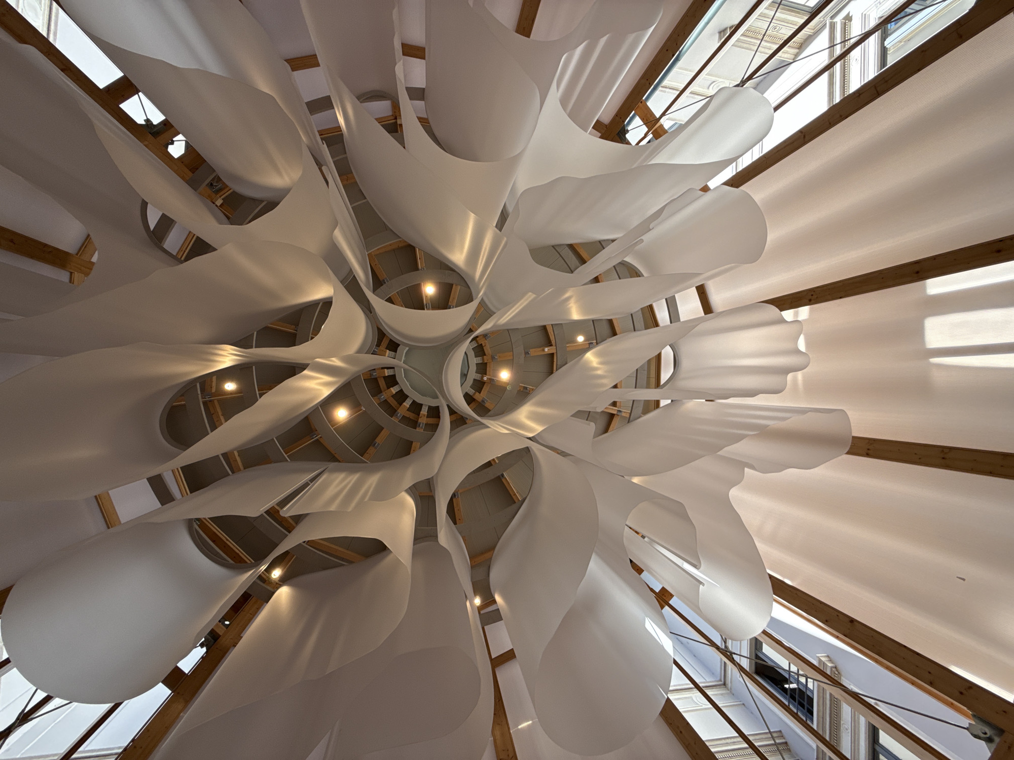 This is a low-angle, wide shot of the interior ceiling of the Fondazione Prada's "Art Club" in Milan, Italy. The ceiling is adorned with large, flowing white fabric panels that are draped and curved, creating a dynamic, sculptural effect. These fabric elements are suspended from a circular wooden and metal frame structure that forms the upper part of the ceiling. Several recessed circular lights are visible within the framework, illuminating the fabric. The overall impression is one of organic, ethereal beauty.