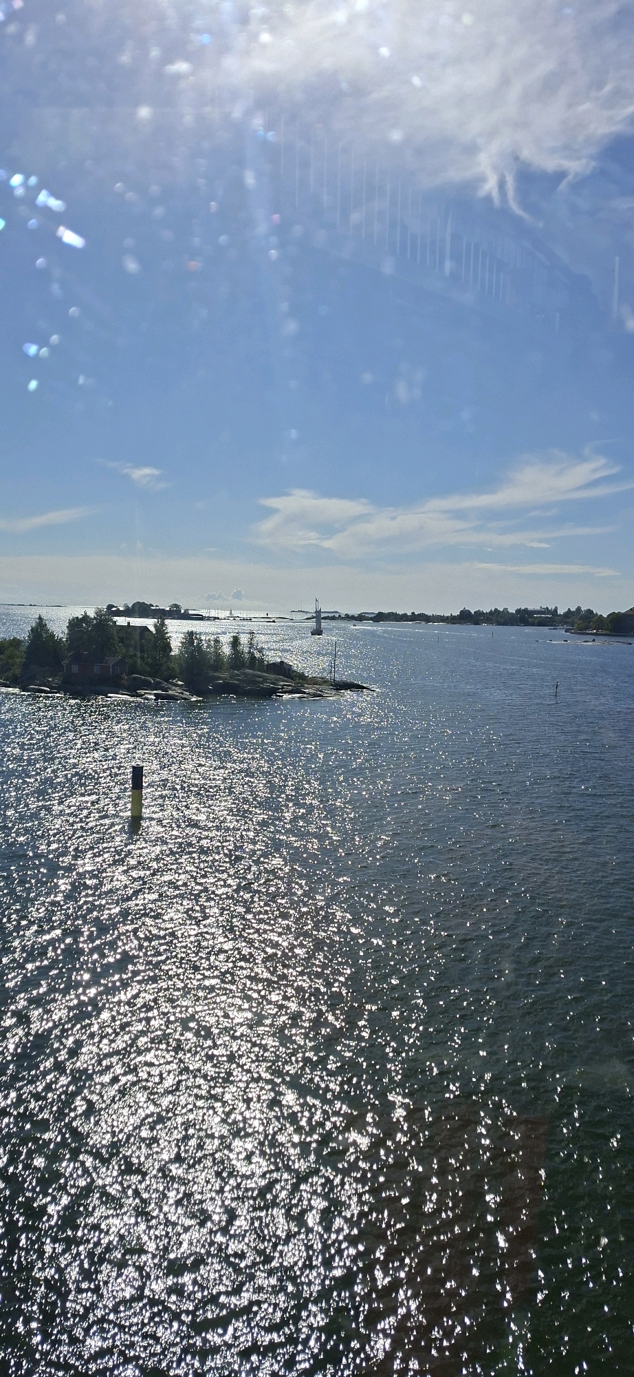 The image is a photograph of a sunny day over the sea in Helsinki, Finland. The foreground features a body of water with strong sun glare reflecting off the surface, creating a shimmering effect. In the mid-ground, to the left, is a small, rocky island with dense green trees and several small buildings, one appearing to be red. A single marker pole, yellow and blue, stands in the water near this island. Further out in the water, a sailboat with a white sail is visible, and in the distance, a hazy horizon line suggests more islands or coastline. The sky is a bright blue with scattered white clouds, and there are some light flares or reflections in the upper portion of the image, potentially from the lens or a window. The scene depicts a calm, natural maritime environment within the archipelago of Helsinki.