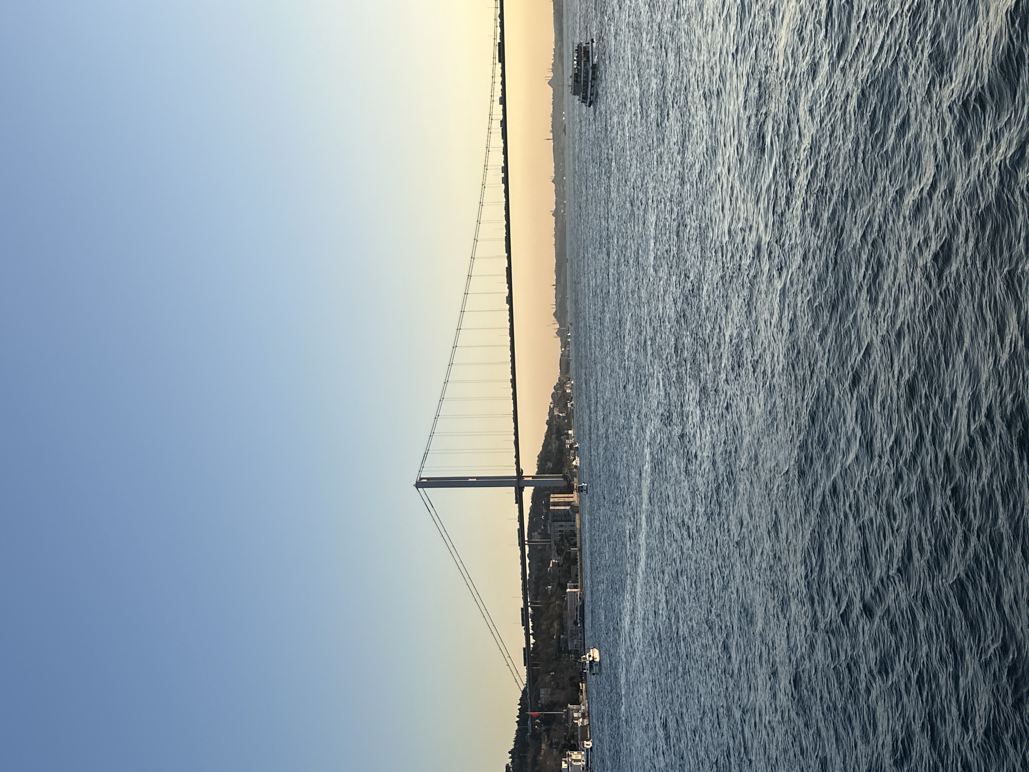 A bridge spans the Bosphorus Strait in Beşiktaş, Turkey. The bridge is a suspension bridge with two prominent towers. Buildings and infrastructure line the strait banks. A boat is on the water in the background. The sky is a gradient of blue to yellow.