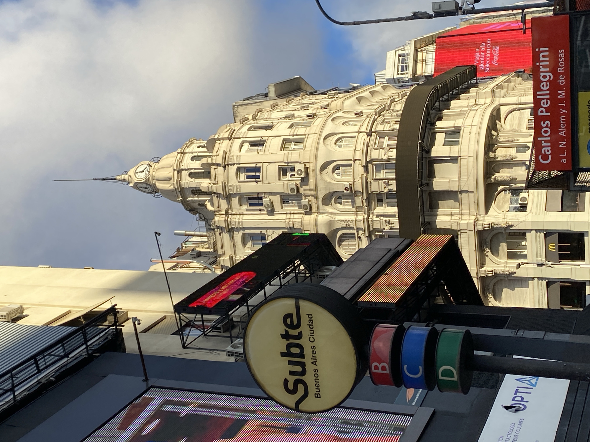 An upward-angled daytime shot captures a vibrant urban scene in Buenos Aires, Argentina. A grand, ornate, light-colored building with a distinctive clock tower and spire dominates the upper portion of the frame, its detailed facade featuring multiple windows and air conditioning units. The sky above is partly cloudy and bright.
In the mid-ground and foreground, a variety of signs indicate a busy commercial and transit area. A prominent circular sign for the "Subte Buenos Aires Ciudad" (subway) is visible, accompanied by colored rings bearing the letters "B," "C," and "D," likely denoting subway lines. Overhead, a large red billboard features text promoting "Viaja a alentar a la Selección con Coca-Cola." A vertical red street sign clearly reads "Carlos Pellegrini" with directions "a L. N. Alem y J. M. de Rosas." Other visible text includes "OPTIA," "TACTOLOGIA," and "TESIS OCULARES." A yellow McDonald's 'M' logo can be spotted on a window of the main building. No direct human activity is depicted; however, the array of signs and the prominent architecture suggest a bustling city environment. The image captures the dynamic atmosphere of a city known for its architectural splendor and public spaces.