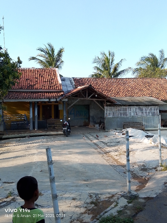 The image depicts a scene in front of a residential building located at coordinates -6.417 latitude and 107.978 longitude in the Kecamatan Anjatan area of Indonesia. The building has a red tiled roof and yellow walls, with a wooden overhang supported by pillars. A motorcycle is parked in the front yard, and there is a partially obscured figure standing near it. The yard is a dirt and concrete surface, with several white poles and a white tarp visible. The background shows two palm trees and a clear blue sky. The scene is likely taken in the afternoon, as the sun is high in the sky and casting shadows.