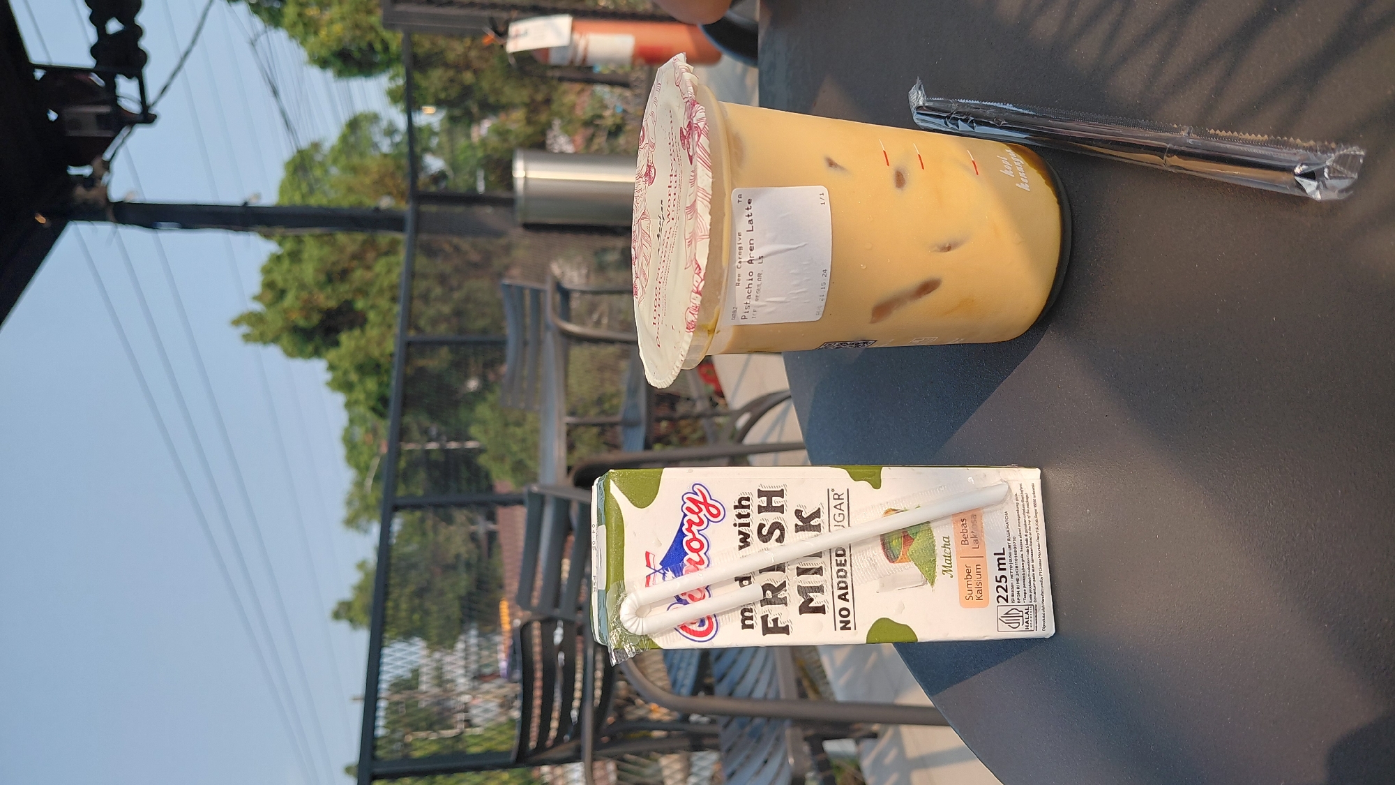 An outdoor cafe scene in Karanglewas, Indonesia, features a table with two drinks and a carton of milk. The table is dark and textured, possibly made of wood or a similar material. In the foreground, a green and white carton of "Greeny" fresh milk with "Matcha" flavor is standing upright. Two white straws are inserted into the carton's opening, one bent at an angle. To the right of the milk carton is a clear plastic cup filled with a light brown, iced beverage, possibly a latte. The cup has a white lid with a red floral pattern and a white sticker that reads "Pistachio Aren Latte." A single black straw, still in its plastic packaging, lies horizontally on the table next to the cup. In the background, out of focus, are outdoor seating areas with chairs and tables, and lush green trees under a clear sky. The overall impression is of a casual, relaxed setting.