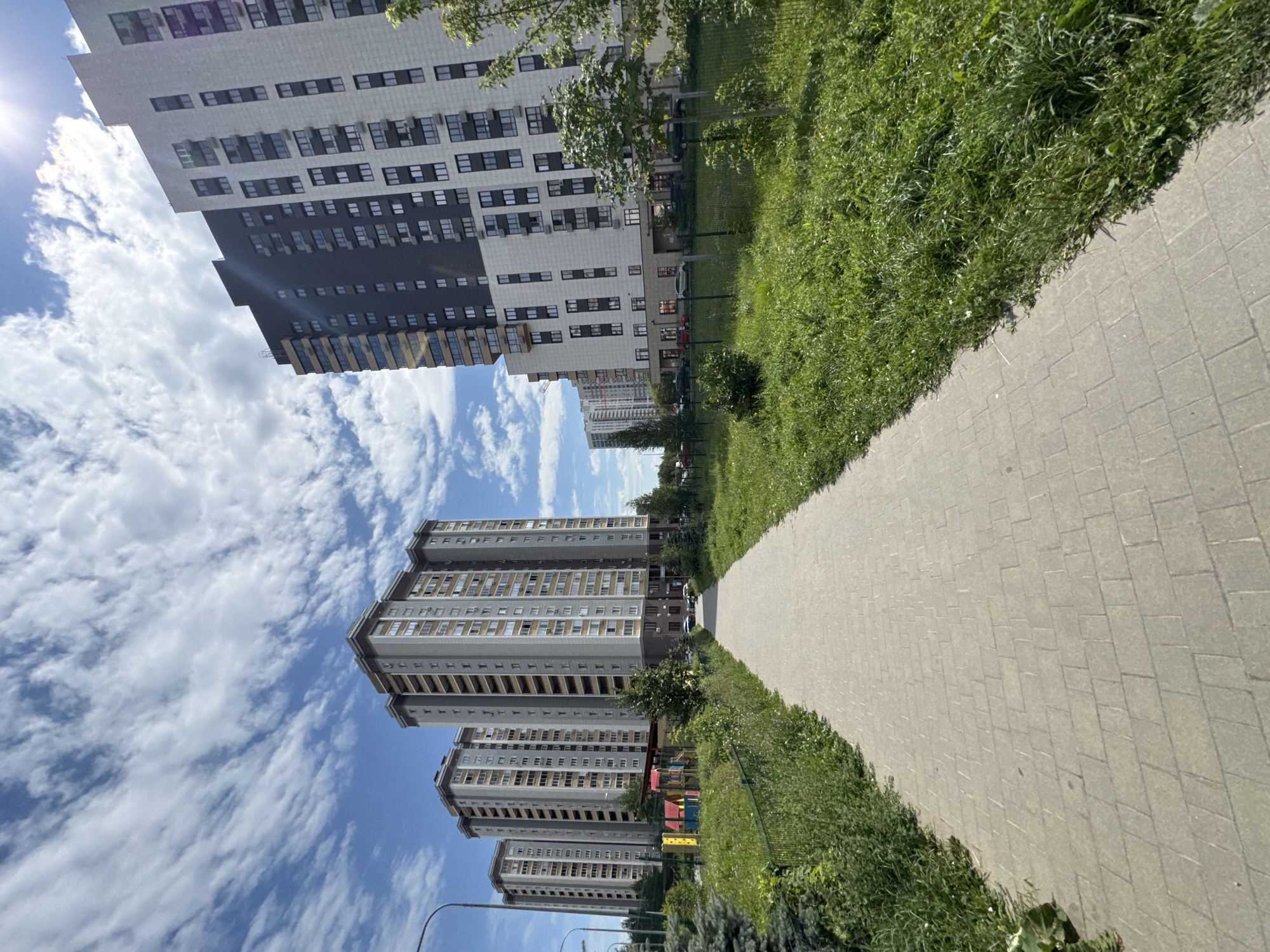 The image shows a wide paved walkway leading towards a cluster of tall, modern apartment buildings. The buildings are of varying designs and heights, with some featuring darker facades and others a lighter, stone-like cladding. Windows are visible on all levels. To the left of the walkway, there is a green fence and a children's playground with colorful structures. Beyond the buildings, more apartment blocks are visible in the distance, suggesting a residential urban area. The sky is bright blue with scattered white clouds, and the sun is shining, creating a lens flare in the upper right corner. Lush green grass and trees line the walkway and surround the buildings. The scene appears to be a well-maintained residential development on a clear, sunny day. This location is in the city of Kazan, Russia.