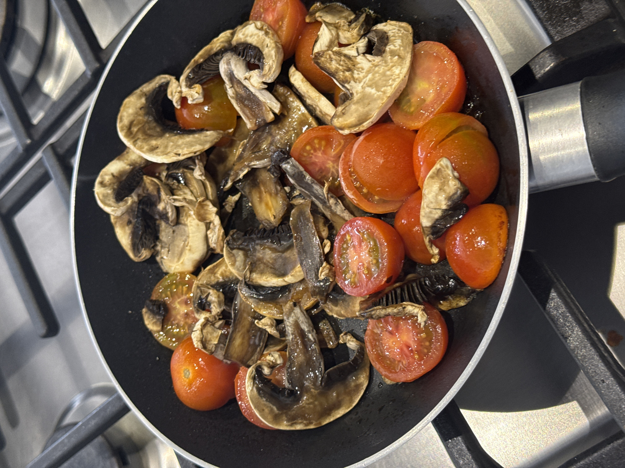 A top-down view shows a black non-stick frying pan sitting on a stainless steel gas stovetop. Inside the pan, sliced button mushrooms and halved cherry tomatoes are sauteing. The mushrooms range in color from light tan to dark brown. The tomatoes are red and glisten with moisture. The stovetop has a grid-like pattern, and the pan handle is visible on the right side. There is no indication of the location being in San Bernardino Tlaxcalancingo, Mexico, or any people or interactions.