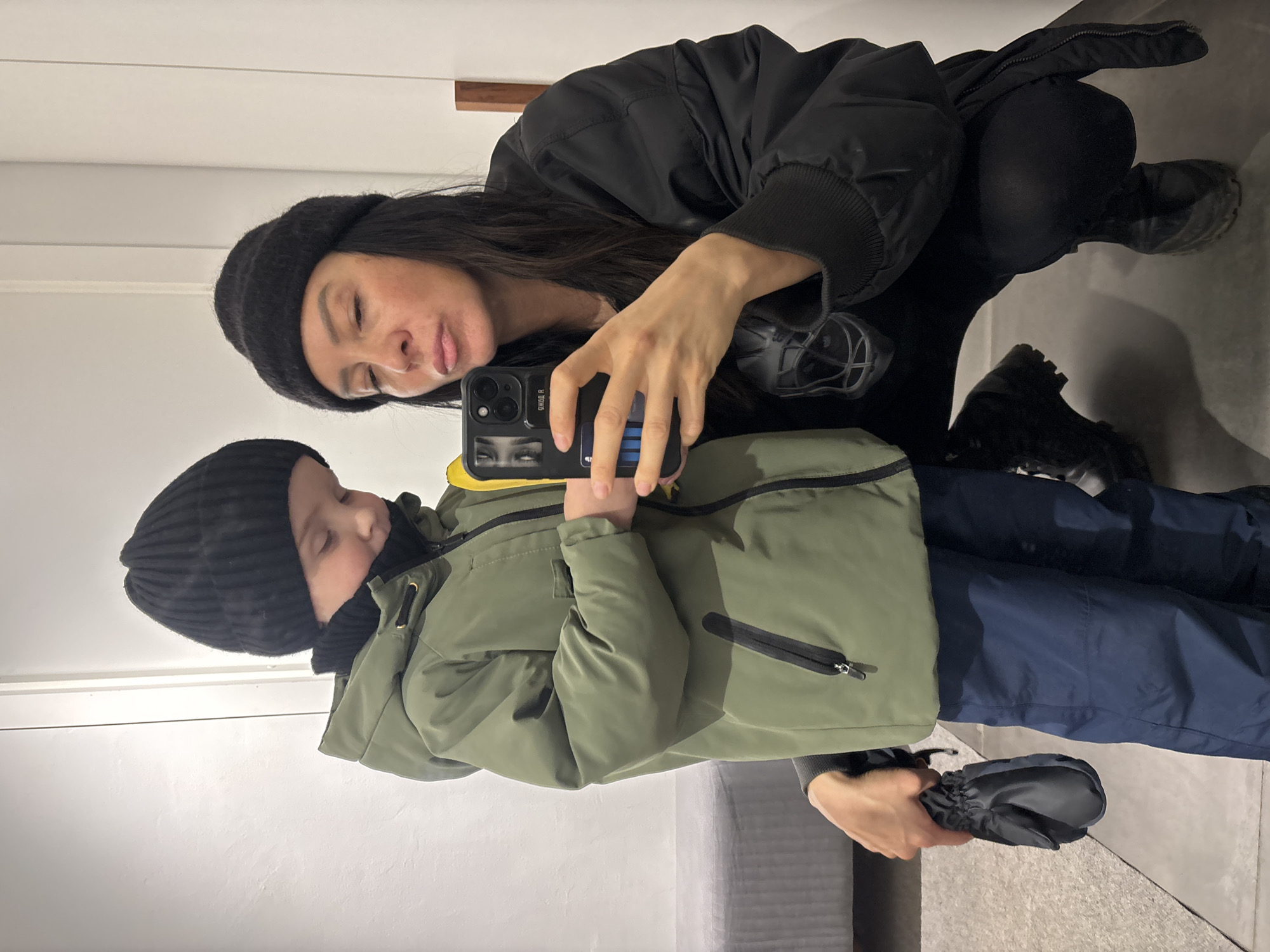 A woman and a child stand close together, both wearing winter clothing, in what appears to be a dressing room or clothing store in Chaikovsky, Russia. The woman, kneeling slightly, wears a black jacket, black beanie, and dark shoes. The child, standing beside her, wears an olive-green winter coat, navy blue pants, a black beanie, and holds a black glove. The woman is taking a photo of them with a smartphone, the phone case displaying an image of eyes. The background consists of a light-colored wall with a thin wooden shelf, a gray padded bench or seat, and a glimpse of the floor.