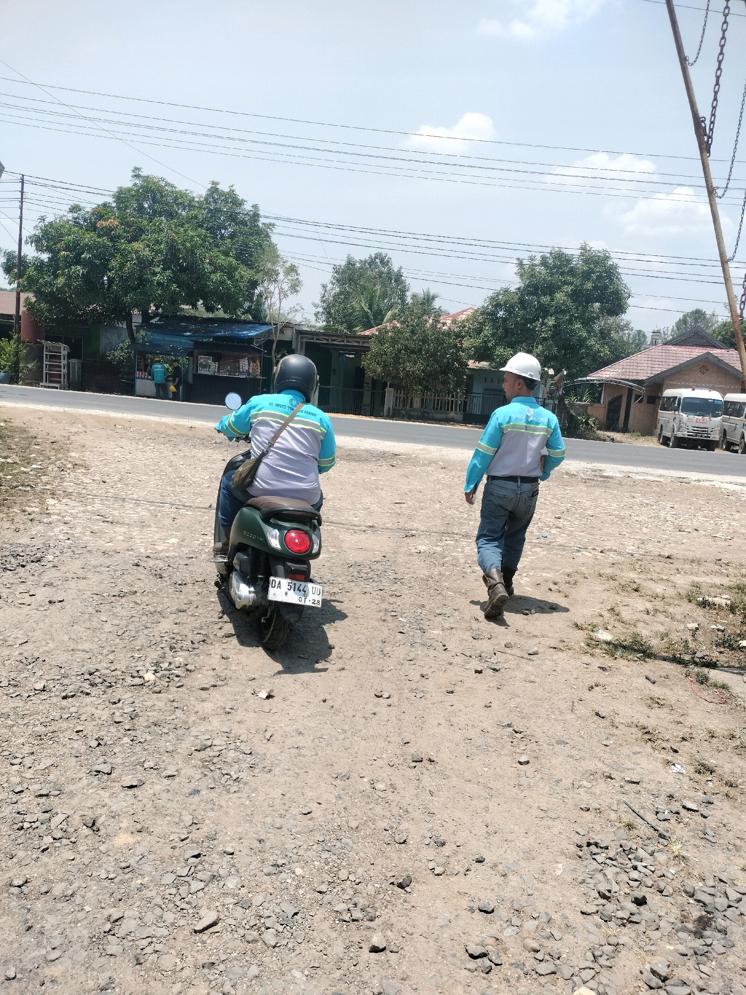 In Maburai, Indonesia, two men are seen in an outdoor, dusty environment, likely on a dirt road or path. The man on the left is riding a dark green scooter and wearing a blue and gray long-sleeved shirt with reflective stripes, a black helmet, and a dark bag slung across his chest. The scooter's license plate reads "DA 5144 UU". The man on the right is walking away from the camera, wearing a similar blue and gray long-sleeved shirt with reflective stripes and a white hard hat. He is also wearing blue jeans and dark boots. Behind them, a paved road is visible with some buildings and trees lining it. A white van is parked on the side of the road. Overhead, power lines are strung across the sky, and the weather appears to be clear and sunny. The ground is covered in gravel and dirt.