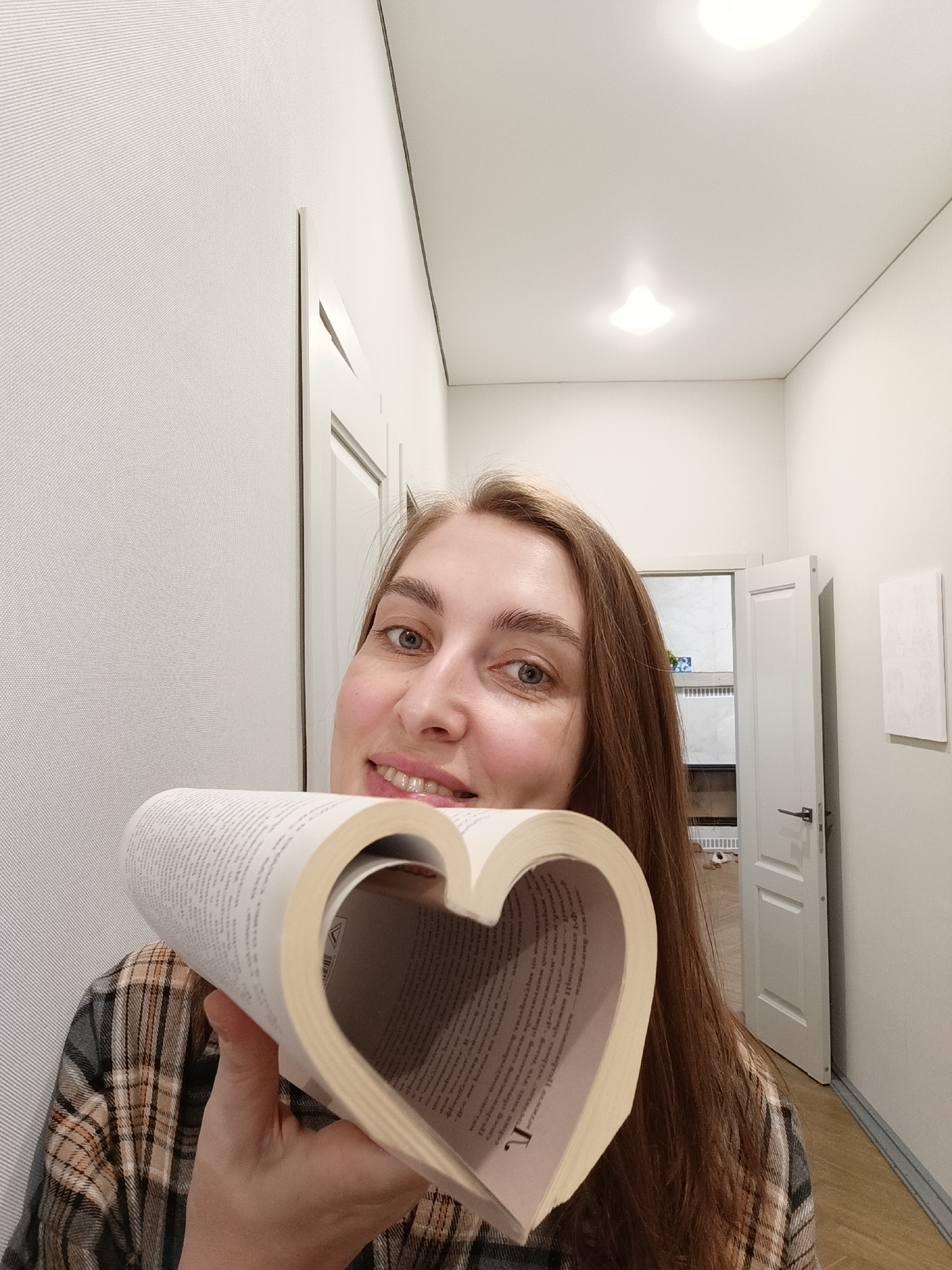 A smiling woman with long, straight brown hair and light blue eyes looks directly at the camera, posing for a photograph indoors. She is wearing a plaid shirt featuring grey, brown, and white patterns. Her hands hold an open book, bending its white pages to form a distinct heart shape. The text printed on the book's pages is largely illegible due to blur and distance, although some letters like "T" are faintly visible in close-ups.
The setting is a brightly lit hallway or corridor with plain, light-colored, subtly textured walls and a matching ceiling. Two recessed square lights in the ceiling provide artificial illumination. In the background, there are two white paneled doors; one on the left is mostly closed, while another on the right is ajar, revealing a glimpse into an adjacent room, possibly a bathroom, with white tiled walls, a white vent, and a dark door handle. A light-colored floor is visible in the lower right corner. The woman's expression is joyful and engaging, suggesting happiness or contentment. The time of day is not discernible due to the indoor artificial lighting. The image was taken in Chaikovsky, Russia.