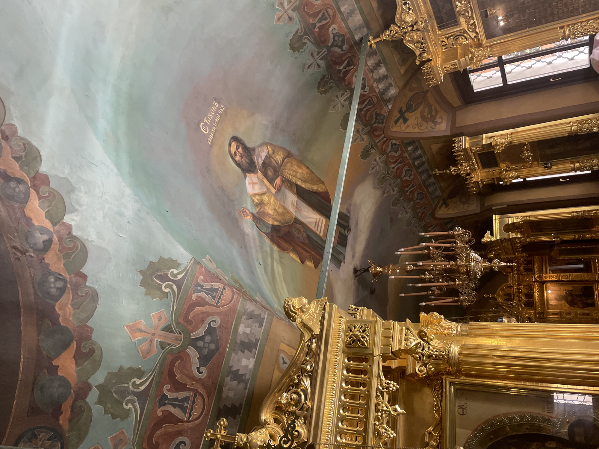 The image shows an interior view, likely inside a Russian Orthodox church in Moscow, Russia, based on the architectural style and religious iconography. The scene is oriented upwards, focusing on a ceiling fresco depicting a male saint, possibly a depiction of Tikhon of Zadonsk (based on the Cyrillic inscription "С Тихомъ" and "Задонский 482."). The fresco is painted in muted blues, greens, and earth tones. The surrounding ceiling is adorned with decorative patterns and cross motifs.
In the foreground, there is ornate gold detailing, part of the iconostasis or other church furnishings. A multi-tiered chandelier with candle-like lights is visible, adding to the atmosphere. A window can be seen in the upper-right portion of the image.