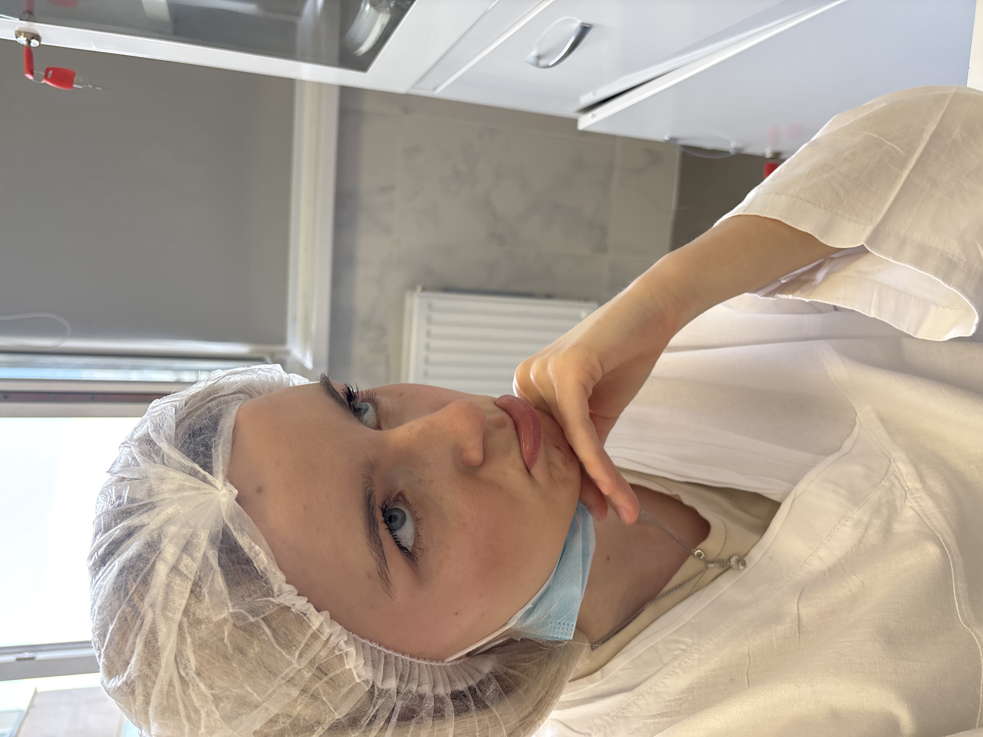 An indoor, tilted medium shot shows a young woman in what appears to be a medical or cosmetic treatment facility, potentially in Ladozhskoe Ozero, Russia. She has fair skin, blue eyes, and light-colored hair, which is covered by a disposable white hair net. A light blue surgical mask is lowered to rest below her chin. She wears a white, loose-fitting smock or gown, under which a lighter-colored top and a thin silver necklace are visible. Her right hand is raised, with fingers gently pressing against her chin and lower lip, lending her a pensive or slightly contemplative expression as she looks upwards and to the left.
The background reveals a clean, clinical environment. Above her are white cabinets, one featuring a small red, key-like attachment. The wall behind her is tiled in a light gray or beige, with a white radiator visible below. A large window to her left provides bright natural light, indicating it is daytime. The scene conveys a sense of waiting or preparation for a procedure in a sterile setting. No legible text is visible in the image.