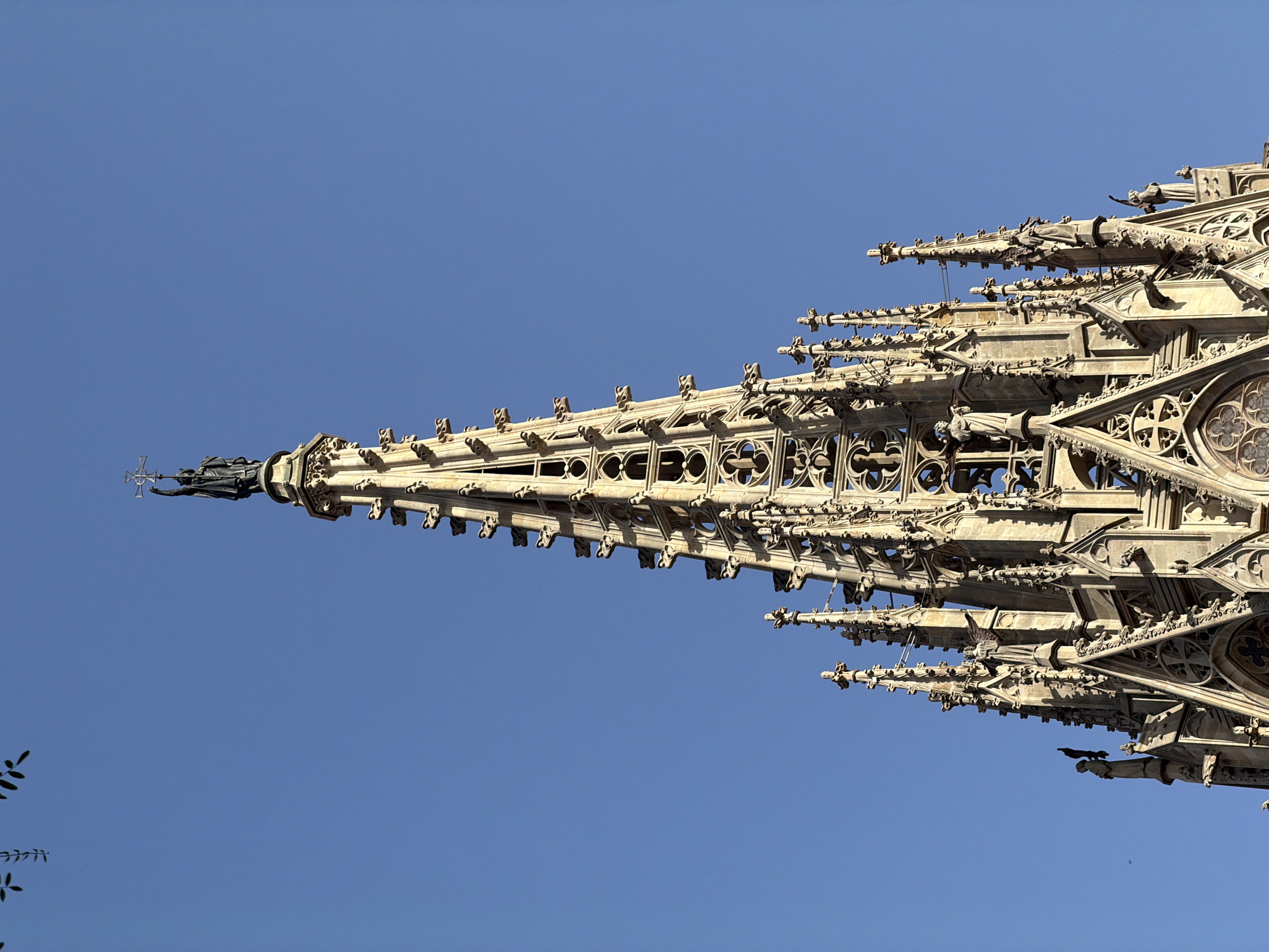 The image shows a close-up of the spire of a Gothic cathedral against a clear blue sky. The spire is elaborately decorated with Gothic architectural elements, including intricate stonework, pointed arches, and numerous small statues and gargoyles. At the very apex of the spire stands a dark-colored statue of a figure holding a cross. The stone of the cathedral appears weathered and light in color. In the upper left corner of the image, a few dark green leaves from a tree are visible. This architectural detail is part of the Sagrada Familia basilica in Barcelona, Spain.