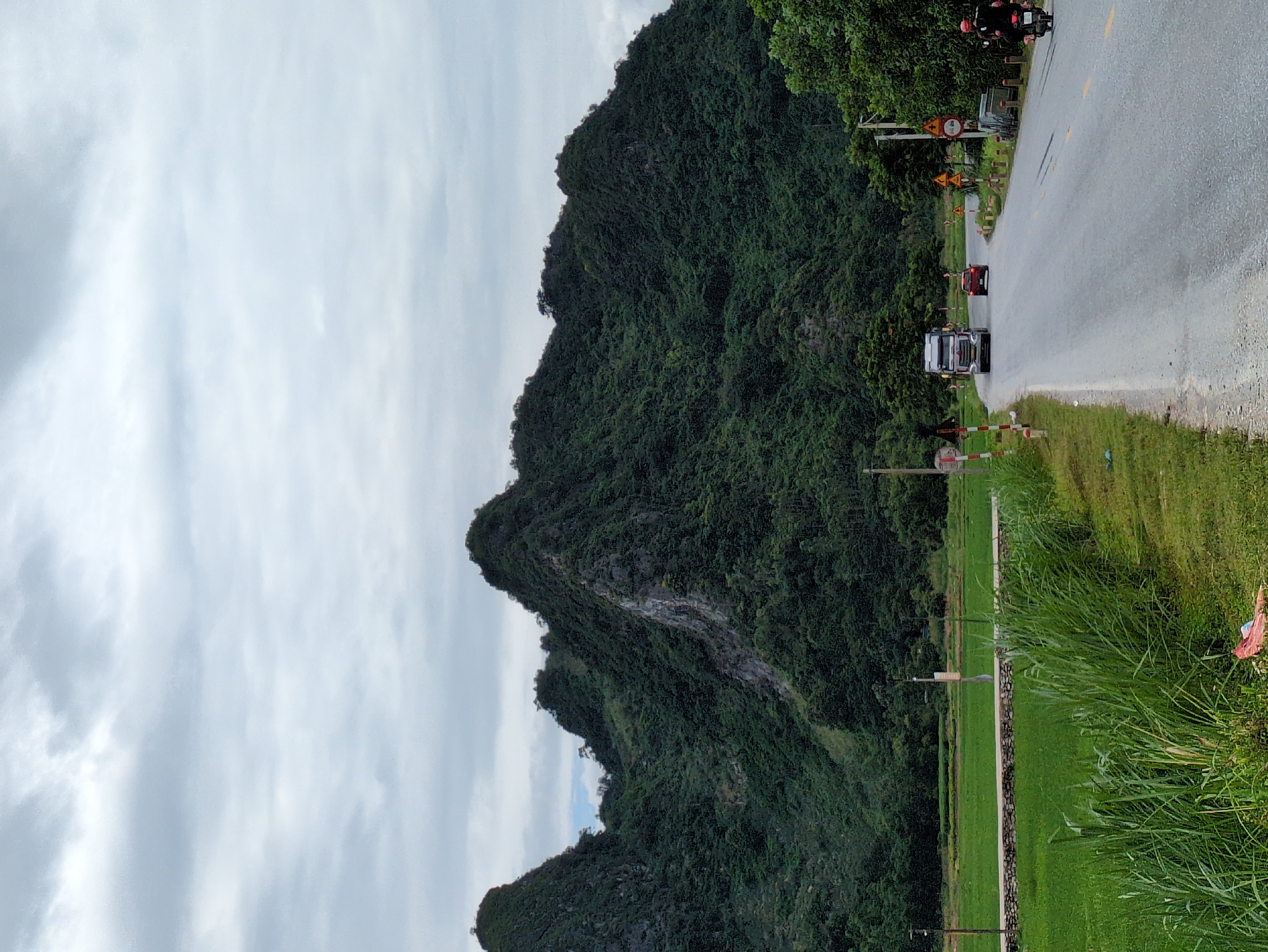 The image depicts a rural road in Tân Thanh Commune, Vietnam, with a prominent, lush green mountain range dominating the background. The sky is overcast with heavy clouds. A paved road curves through the scene, with a truck and a red car traveling on it. On the right side of the road, a person is riding a motorcycle. The left side of the road features a vibrant green rice field bordered by a stone wall and tall grass. Several traffic signs and poles are visible along the roadside. The overall impression is one of a scenic, agricultural landscape.