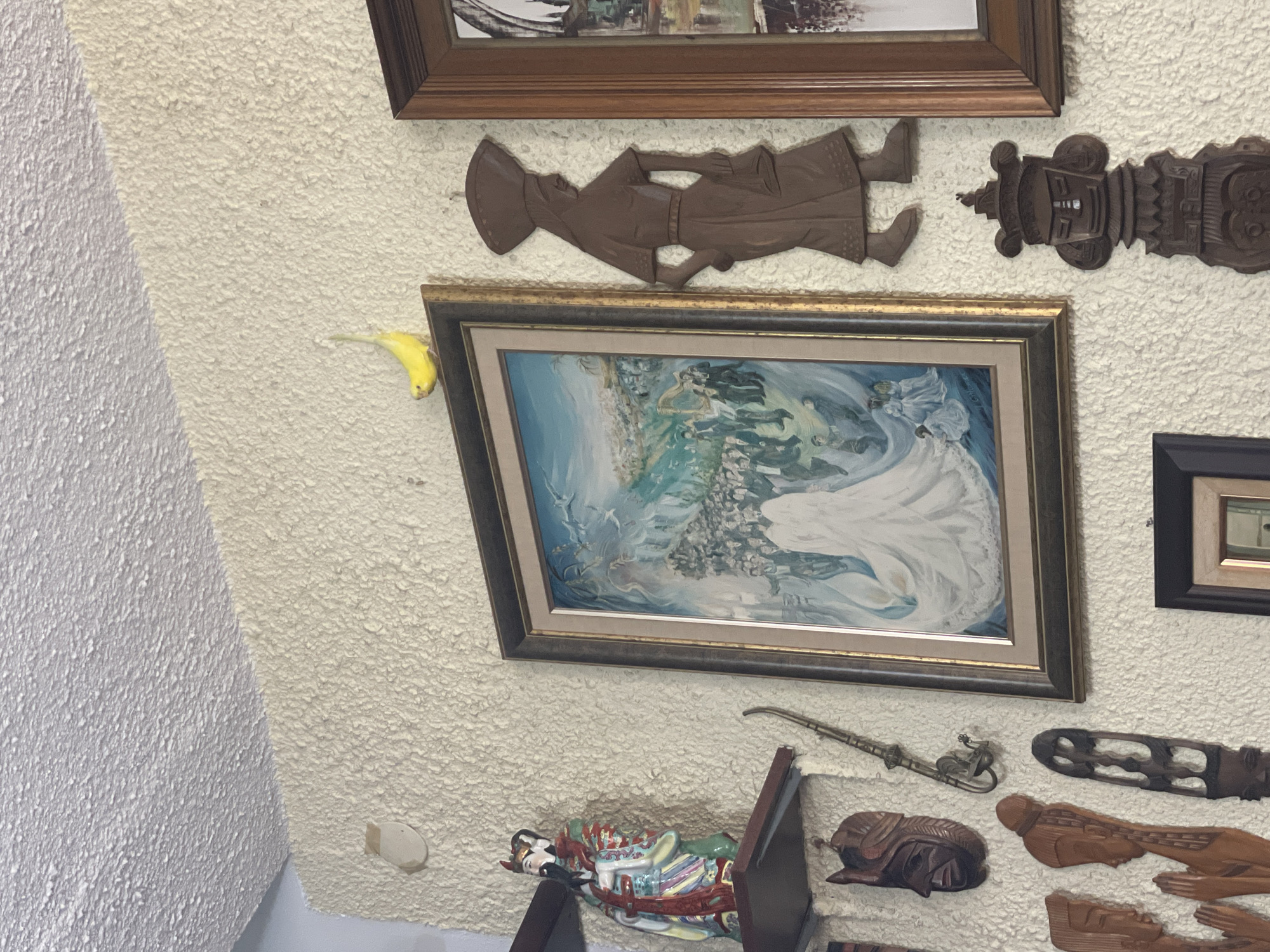 The image shows a section of a textured white wall. On the wall are various decorative items, including multiple framed pictures, wooden carvings, and other small figurines or ornaments. A yellow banana is positioned to the left of a painting. The angle of the shot suggests the wall may be angled or part of an upper floor area. It is not possible to confirm the location as Petah Tikva, Israel from the provided image.