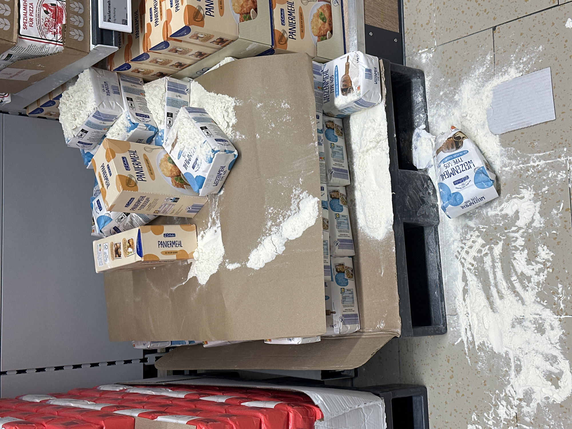 The image shows a spill of flour and related products, possibly in a retail setting, likely a grocery store, in Jestetten, Germany. Several packages of "Weizenmehl Type 405" (wheat flour type 405) and "Paniermehl" (bread crumbs) are visible, some with damaged packaging and flour scattered around them. One bag of "Weizenmehl" is torn and lying on the floor with a significant amount of flour spilled around it. The spill is on a tiled floor next to a black plastic pallet. The products appear to be on shelves or displays in a retail environment. Other products like "Spezialmehl Fur Pizza" are visible on a shelf above.