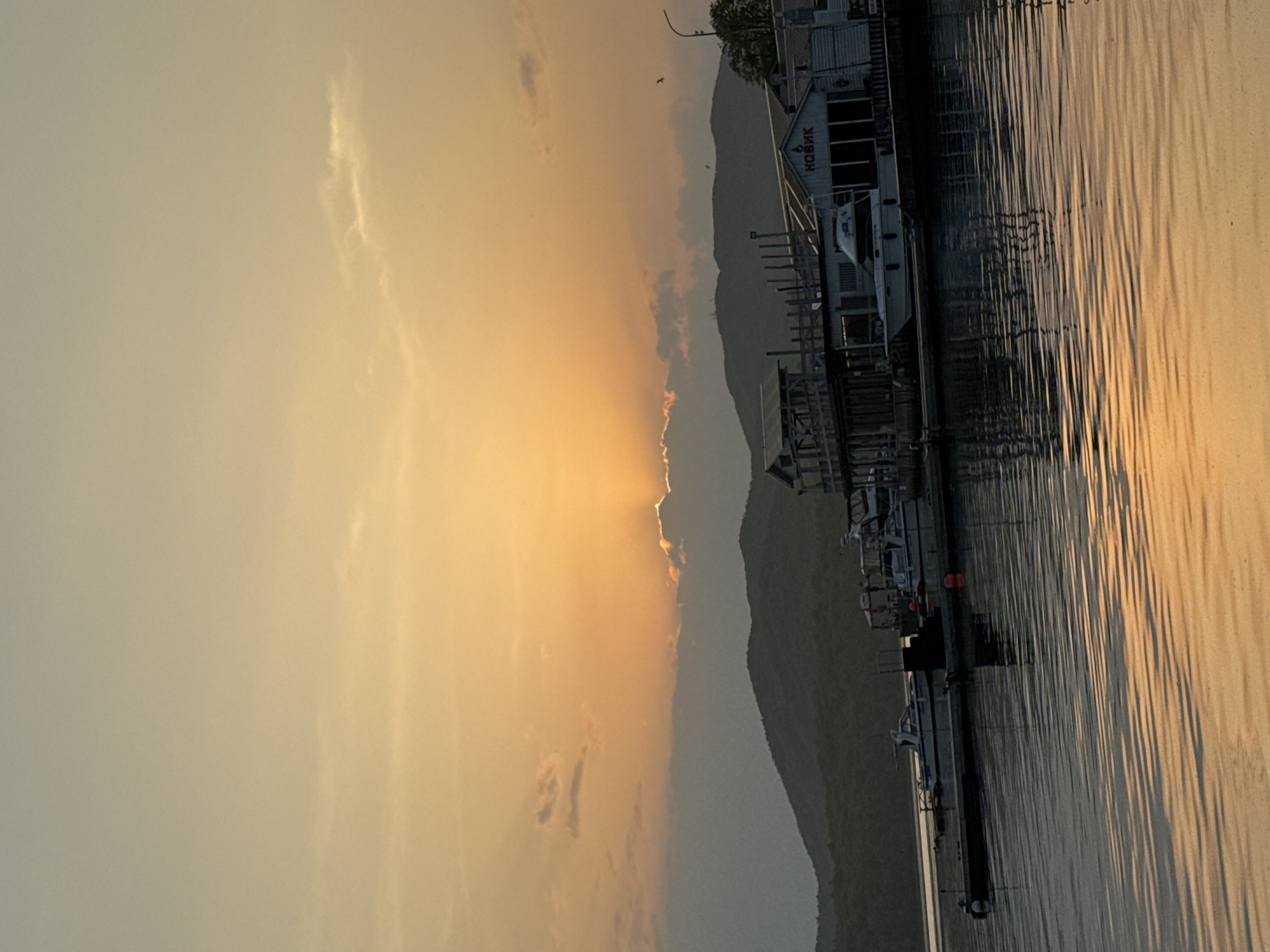 A landscape shot showing a body of water, possibly a bay or harbor, reflecting a sunset. The sun is setting behind a mountainous coastline. On the right side of the frame, there is a dock structure with several boats moored alongside it. A building with "Hobuk" or "KOBMK" written on the front is also visible on the dock. The sky is a mix of yellow and orange hues, indicating the sunset. The photo appears to be taken in the city of Russky, Russia.