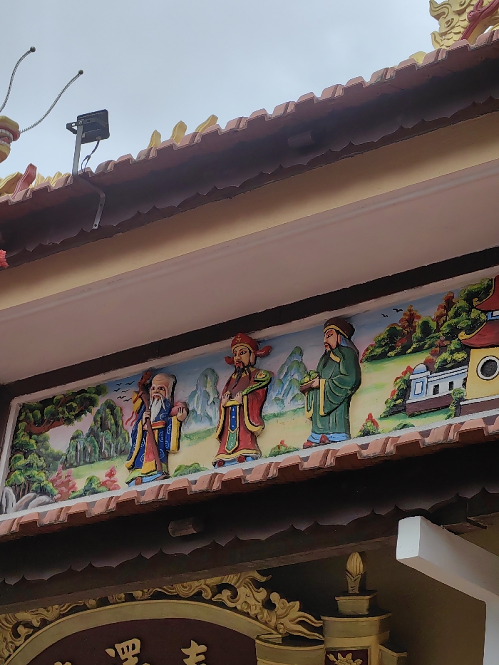 This image captures a detail of a building facade, likely a temple or pagoda, in Phú Xuân Ward, Vietnam. The scene is dominated by a vibrant, rectangular relief panel featuring three anthropomorphic figures, commonly identified as the Fú Lù Shòu (Three Star Gods).
From left to right:
The first figure is an elderly male with a long white beard and a prominent forehead, dressed in a blue robe with yellow trim. He holds a staff in his left hand and an indeterminate object in his right.
The central figure is a male with a dark beard, wearing a red robe with intricate yellow and blue patterns and a red cap. He holds a green ruyi scepter in his right hand.
The third figure is a male with a dark beard, clad in a green robe with yellow trim and a dark cap. He holds a cluster of peaches in both hands.
The background of the relief depicts a stylized landscape with green mountains, trees, and small, traditional buildings under a light blue sky with several bird silhouettes.
Above the relief, a row of reddish-brown, undulating roof tiles is visible, surmounted by a lighter-colored eave. A black rectangular floodlight or surveillance camera is mounted on a light-colored metal bracket on the roof, with visible wiring extending from it. To the immediate left of the light, a yellow and red decorative element, possibly part of a dragon or mythical creature, with two spring-like wires protruding, is partially visible.
Below the relief, an ornate dark brown frieze features golden decorative patterns and partially visible Vietnamese script, forming part of a larger inscription or sign. Beneath this, a plain light-colored (possibly pinkish-beige) wall surface is present. The sky above the building appears overcast and light grey.