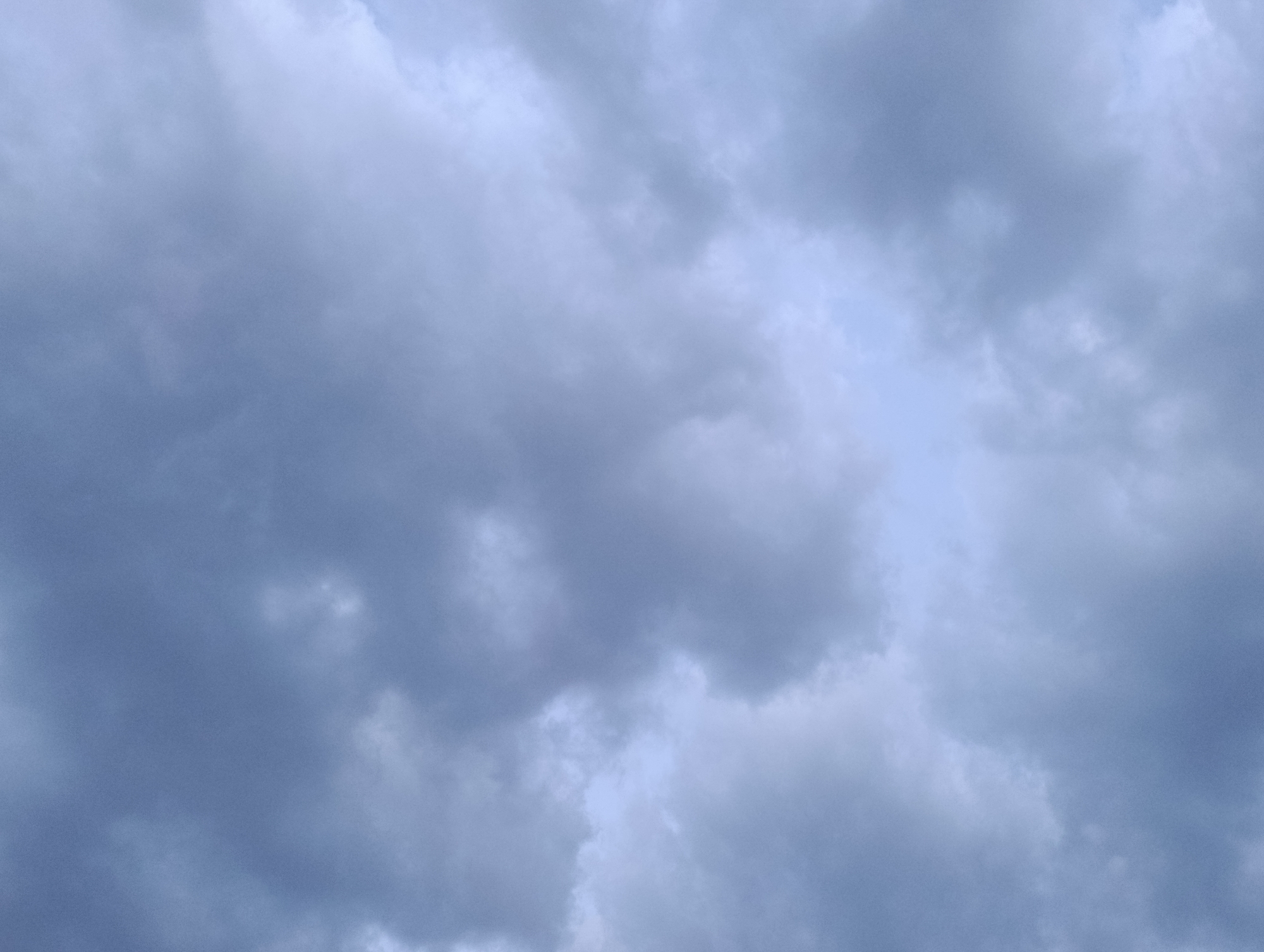 The image is a close-up, low-angle shot of a cloudy sky. The clouds are predominantly a dark, overcast gray, with some lighter patches of white and pale blue suggesting the presence of sunlight filtering through. The texture of the clouds varies from wispy and thin to dense and voluminous. There are no people, structures, or any other objects visible in the frame, as the view is entirely focused on the sky. The lighting is diffused, creating a somewhat muted and somber atmosphere. The scene is entirely natural and appears to be an outdoor environment, but no specific location within Manghit, Uzbekistan can be identified from the visual information provided.